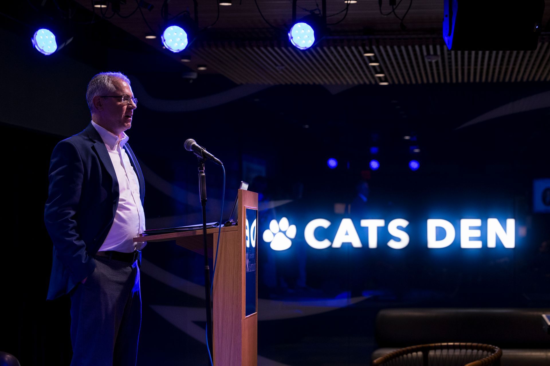 Man standing at a podium on a dim stage beside a glowing “CATS DEN” sign