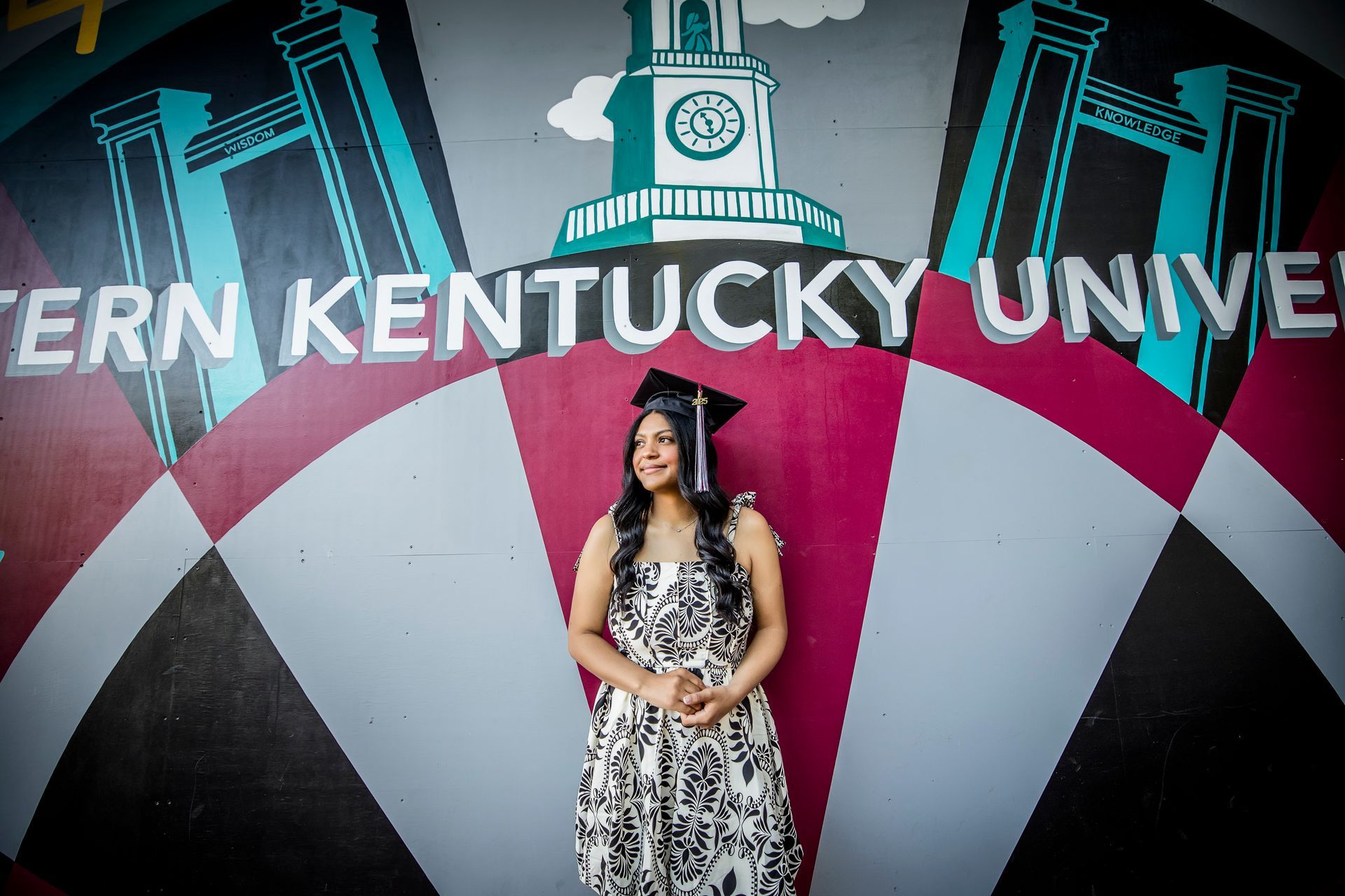 Woman in a graduation cap and dress standing before a Western Kentucky University mural