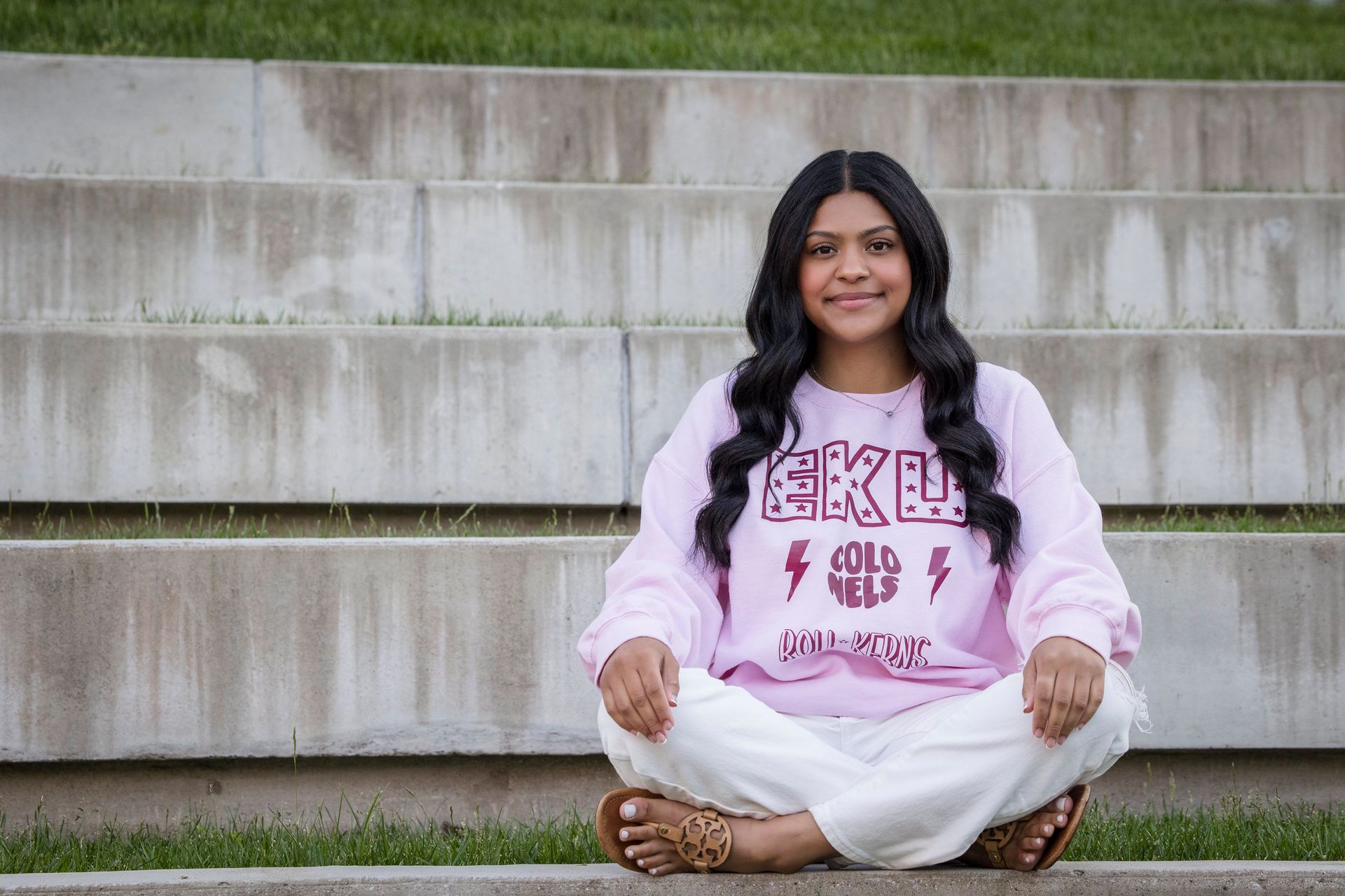 Person sitting cross-legged on concrete steps, wearing a pink sweatshirt and white pants, smiling at the camera.