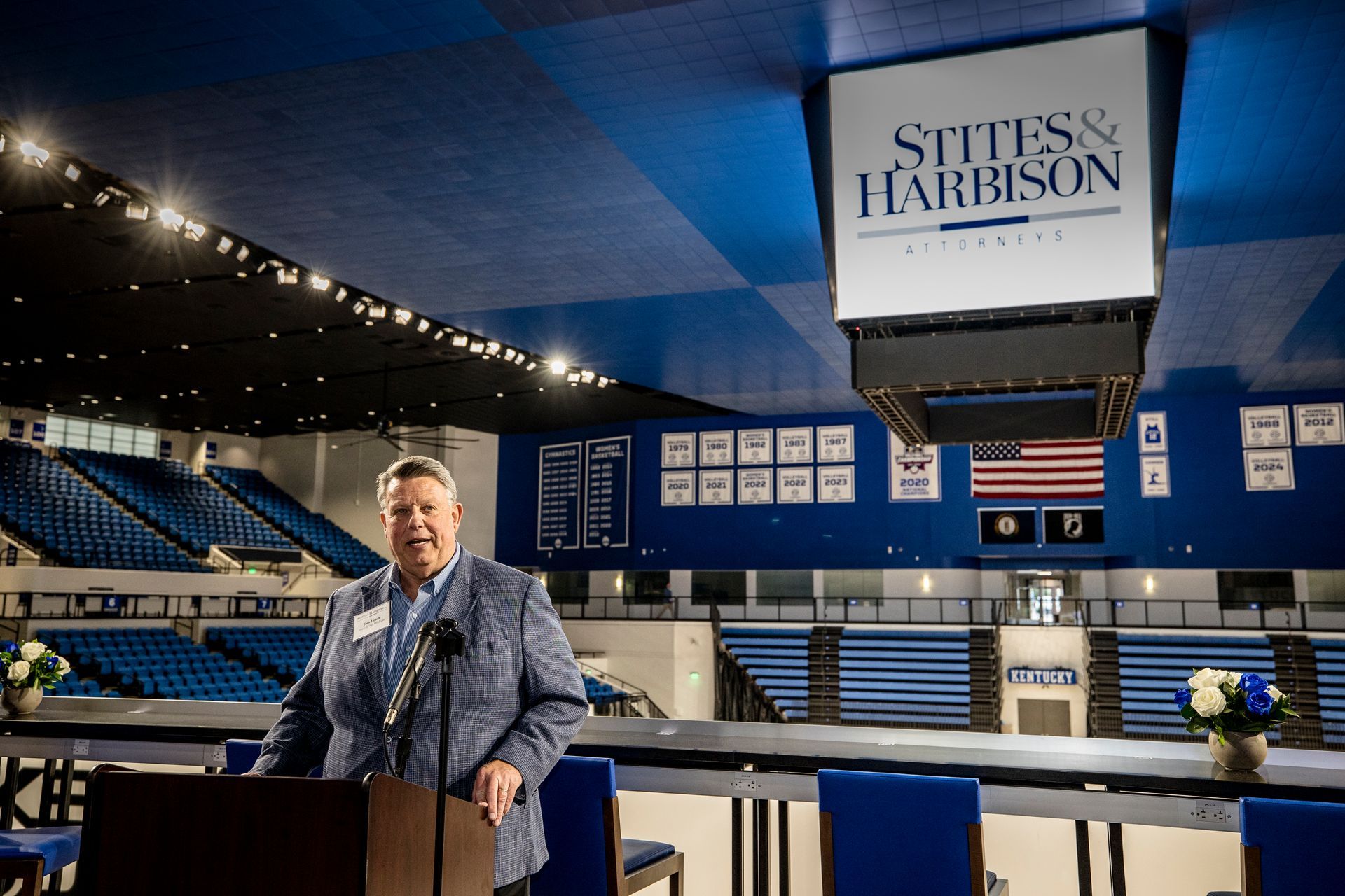 Man speaking at a podium in an indoor arena with blue seating and a large “Stites & Harrison” sign above.