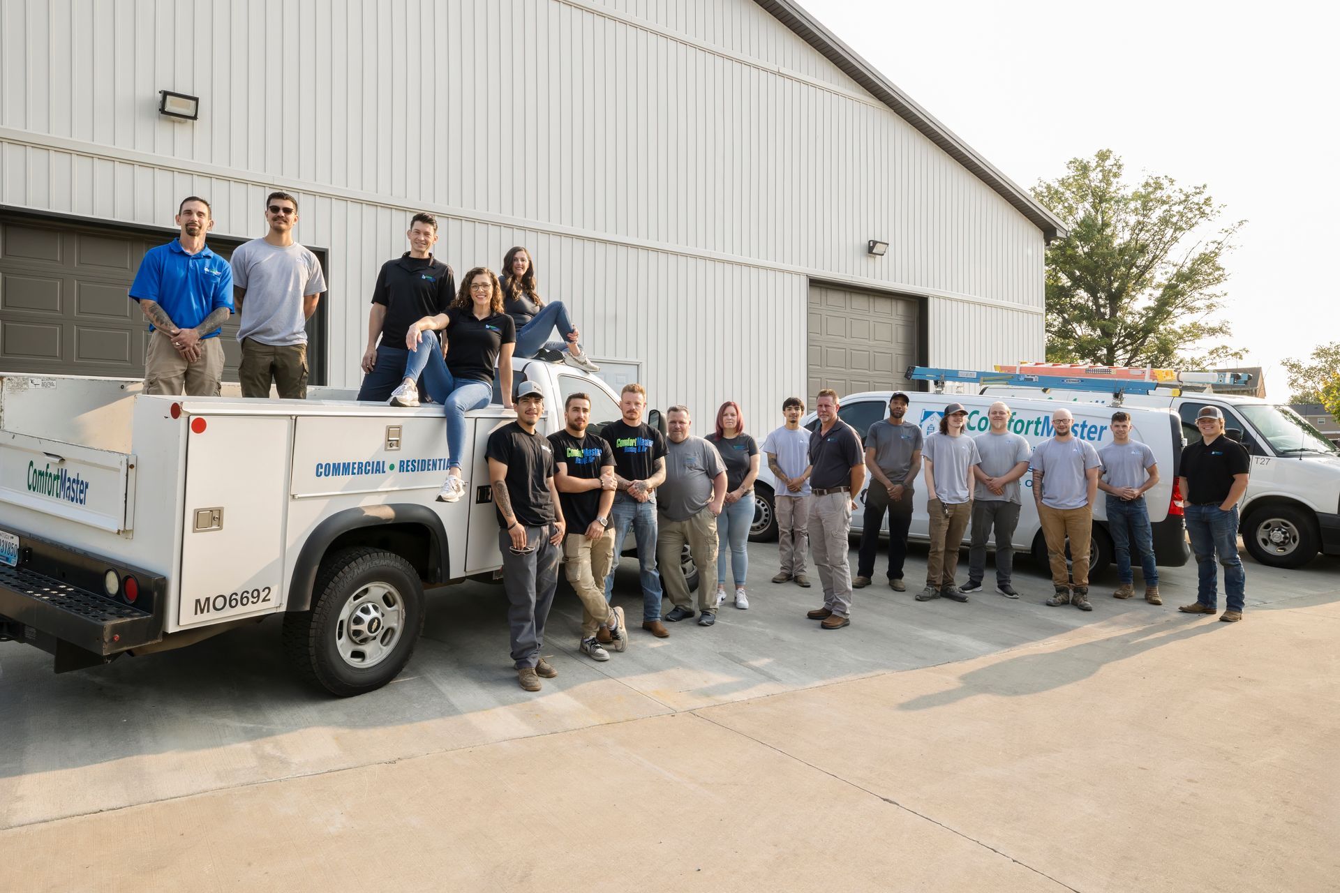 Group of people posing beside a white utility truck outside a warehouse under bright sunlight