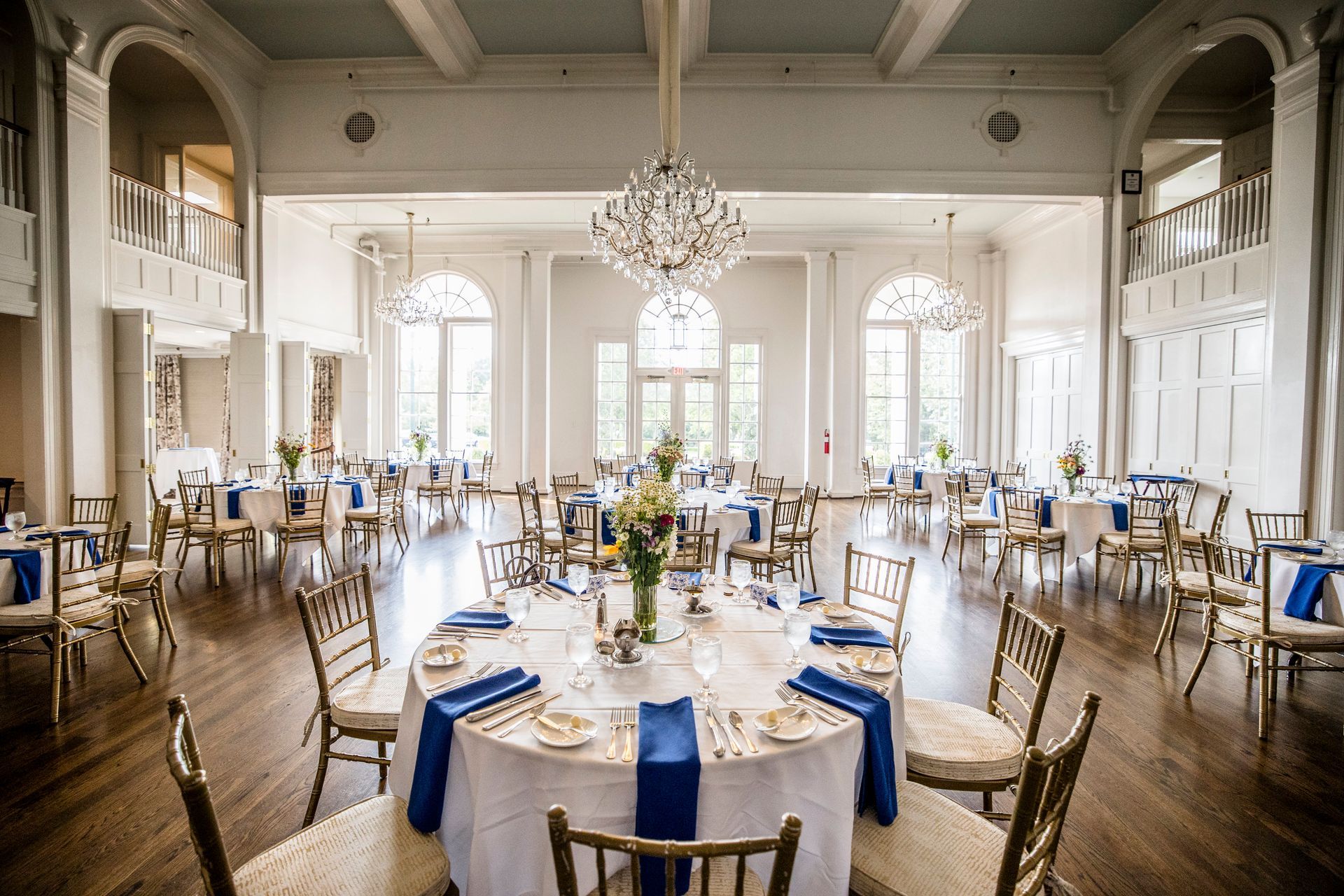 Elegant banquet hall with round tables, white chairs, blue napkins, and chandeliers in a bright room
