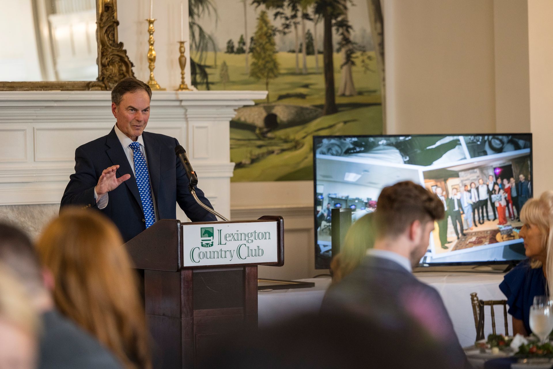 Man speaking at a podium in a formal room, with a presentation screen showing a seated audience and slides