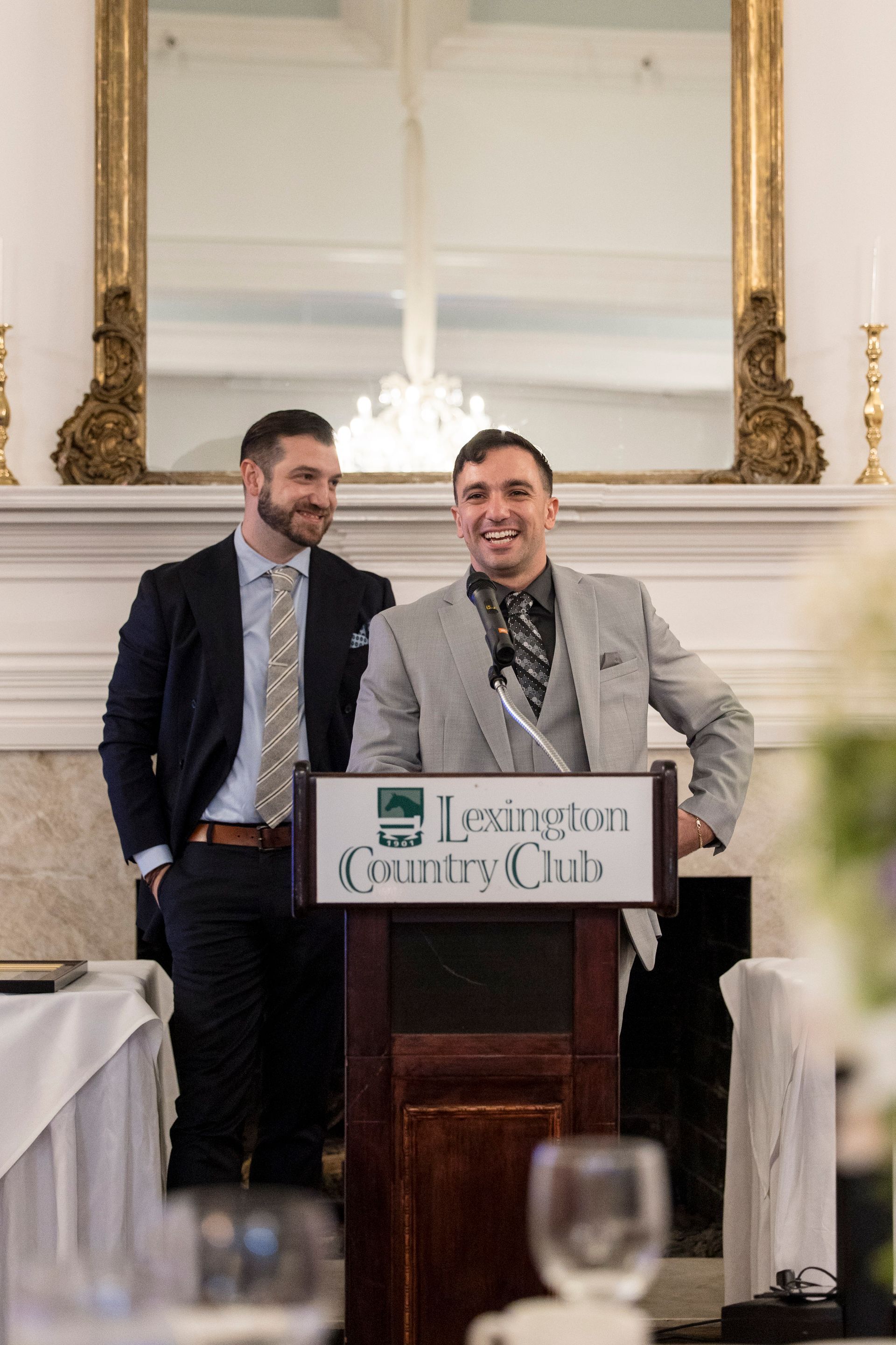 Two men speak at a podium labeled “Lexington Country Club” in a formal banquet hall.