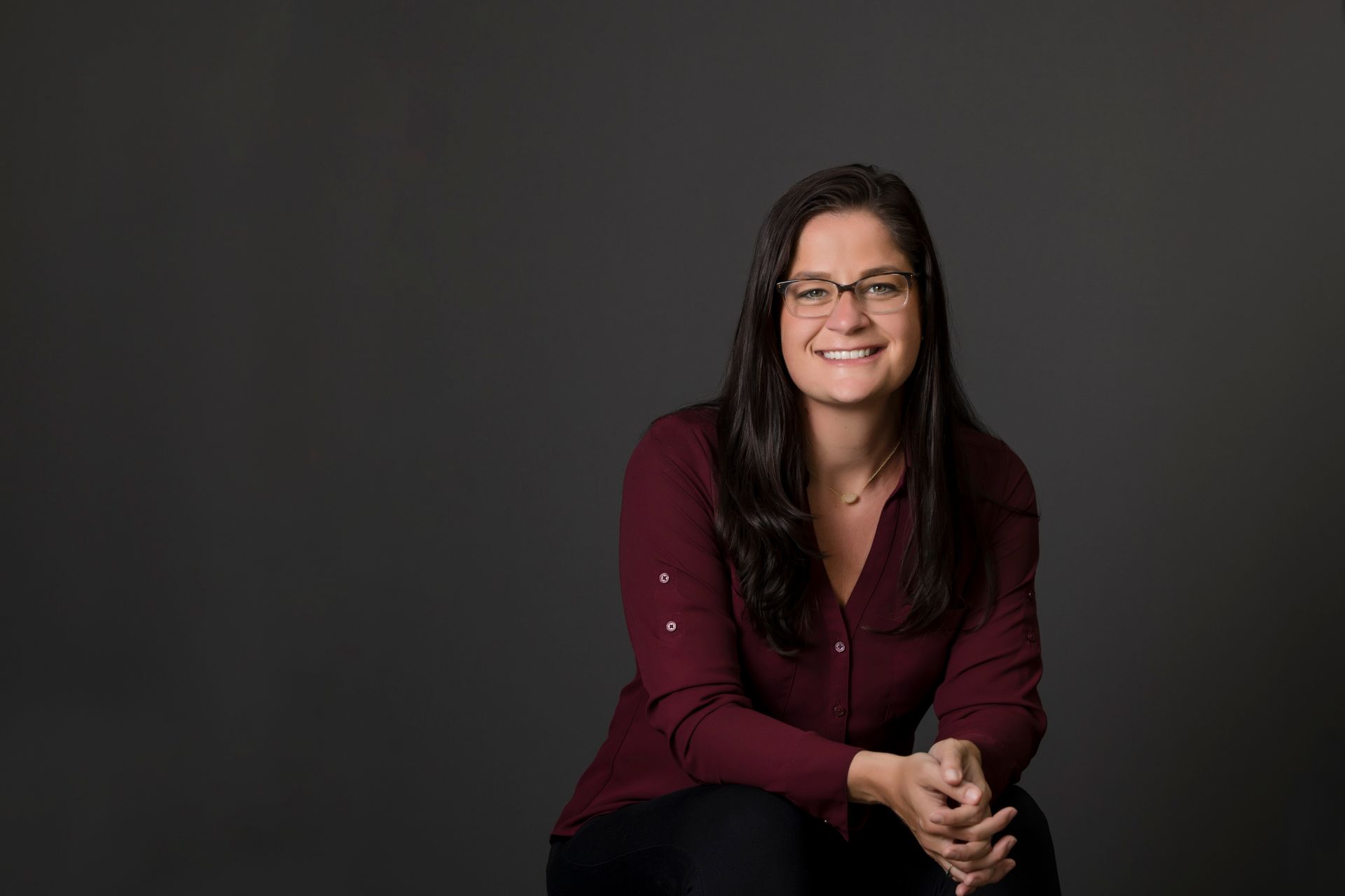 Smiling woman seated against a dark gray background, wearing a burgundy blouse and black pants.