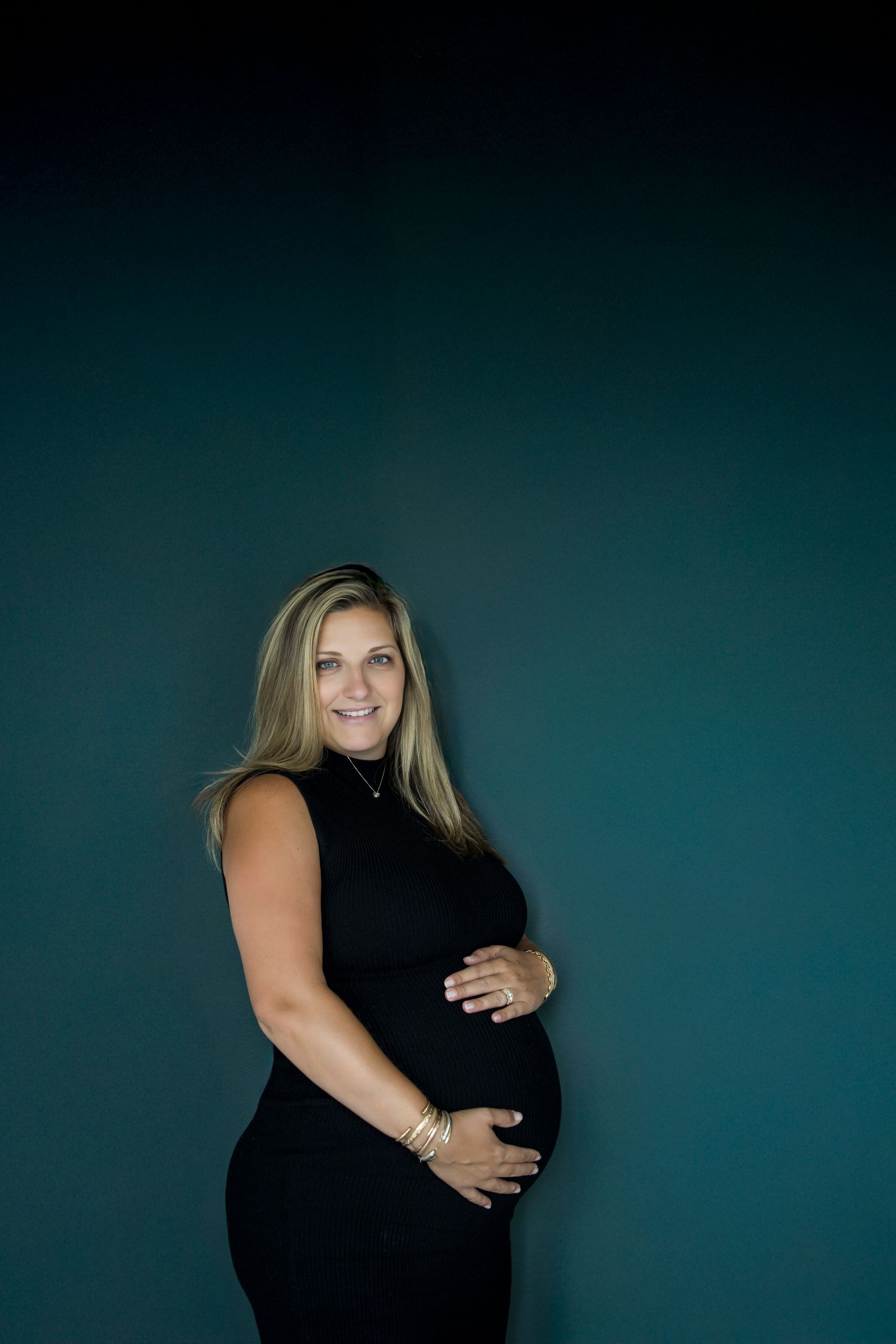 Smiling pregnant woman in a black sleeveless dress standing against a teal background