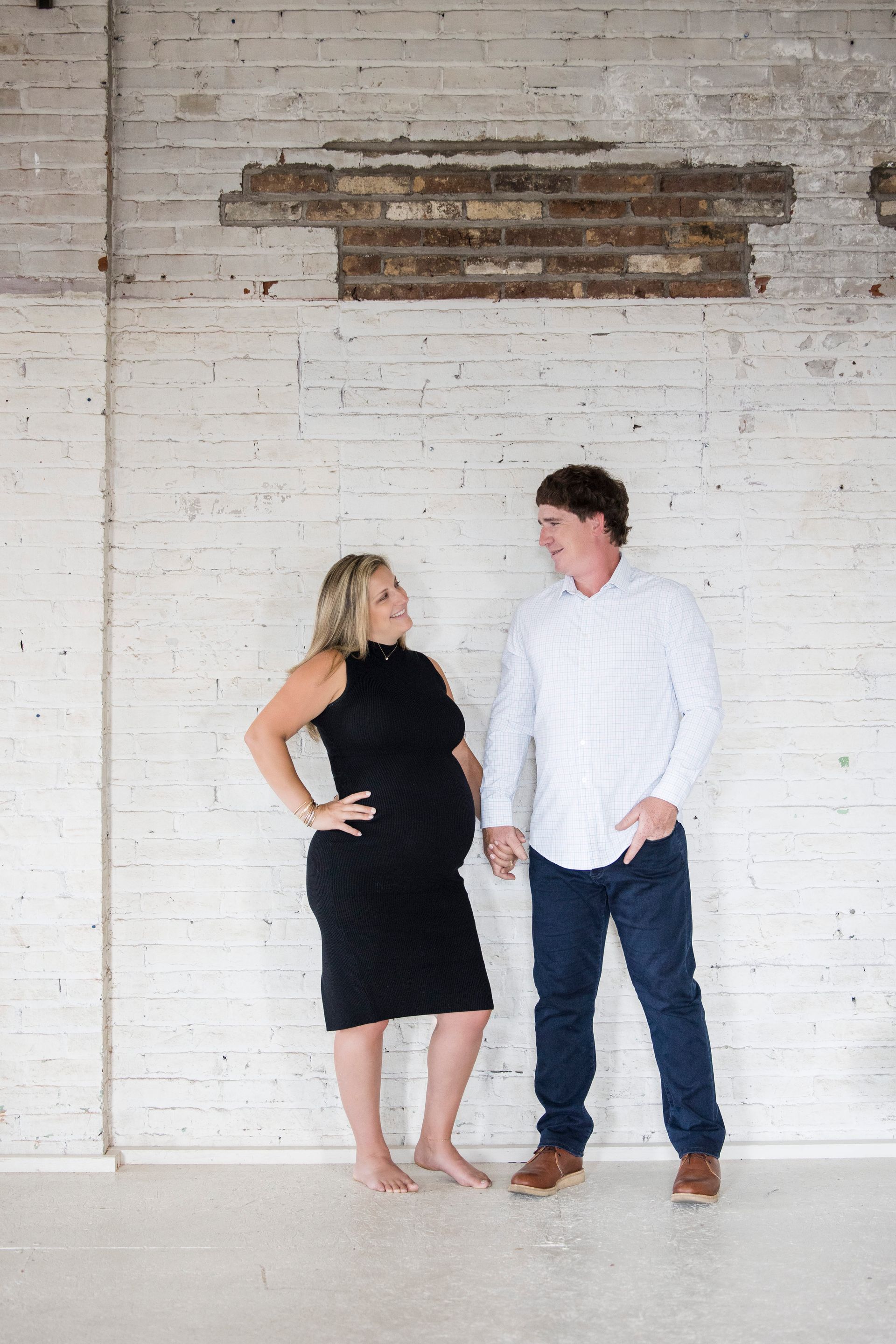 Two people standing and facing each other against a white brick wall, one in a black dress and one in a white shirt.