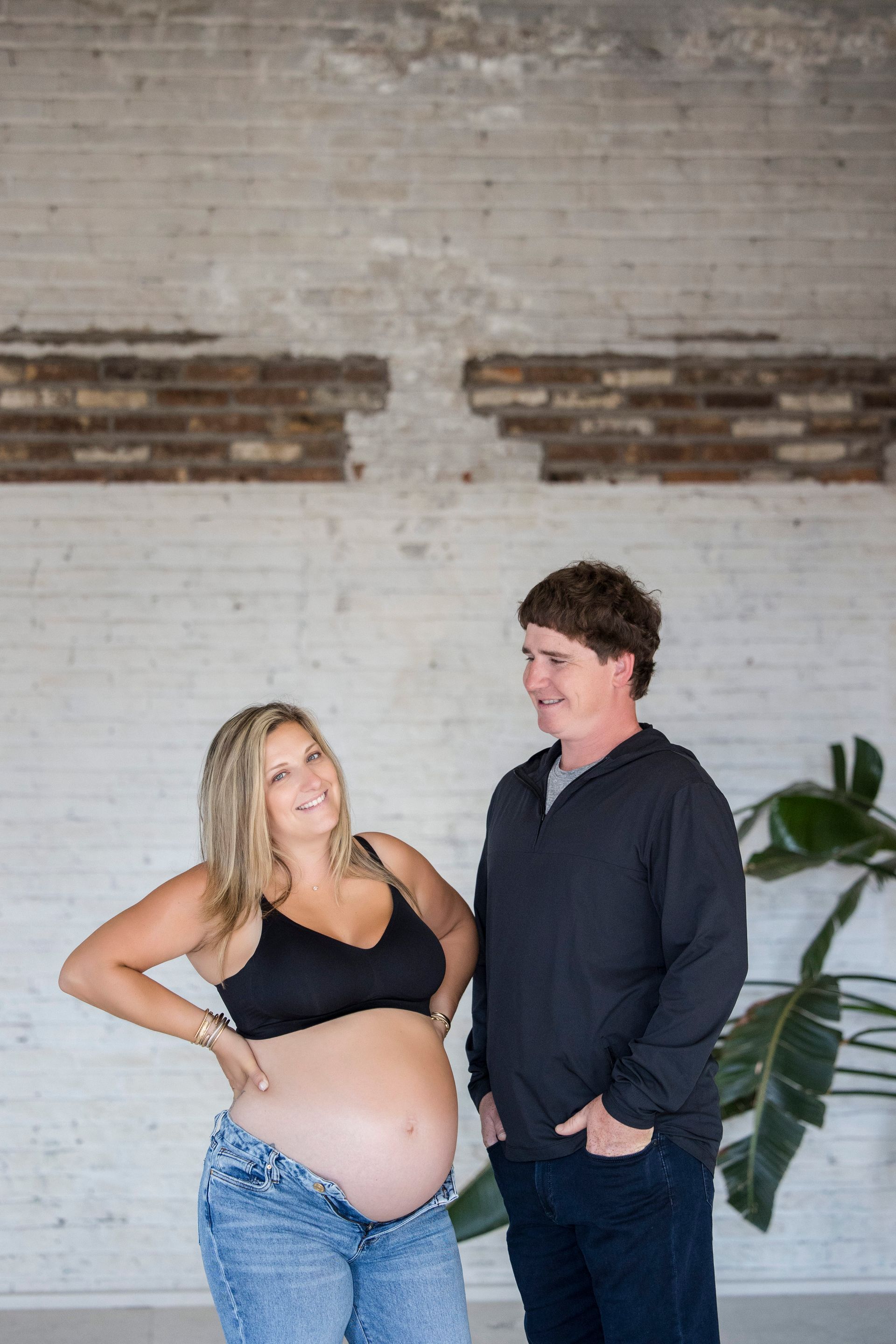 Pregnant woman and man standing together indoors, smiling against a white brick wall.