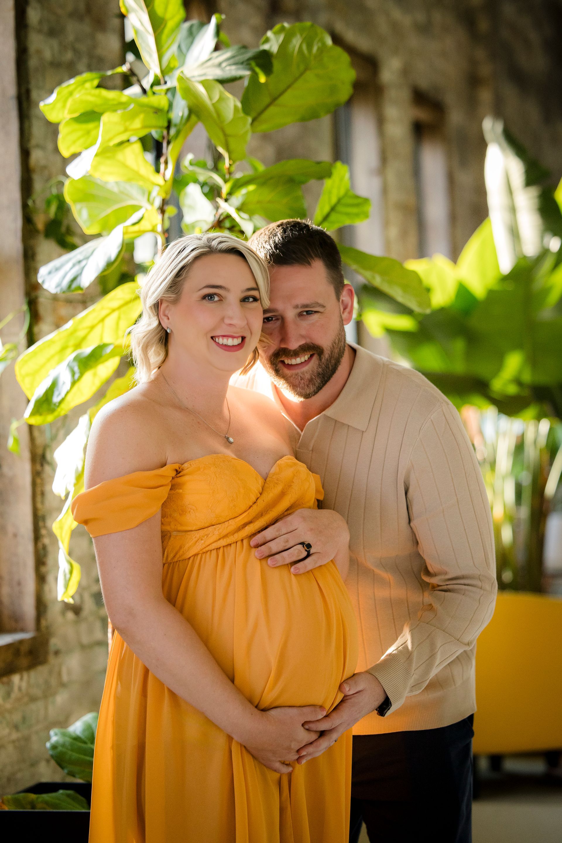 Couple smiling outdoors, with the pregnant woman in a yellow dress and man standing behind her.