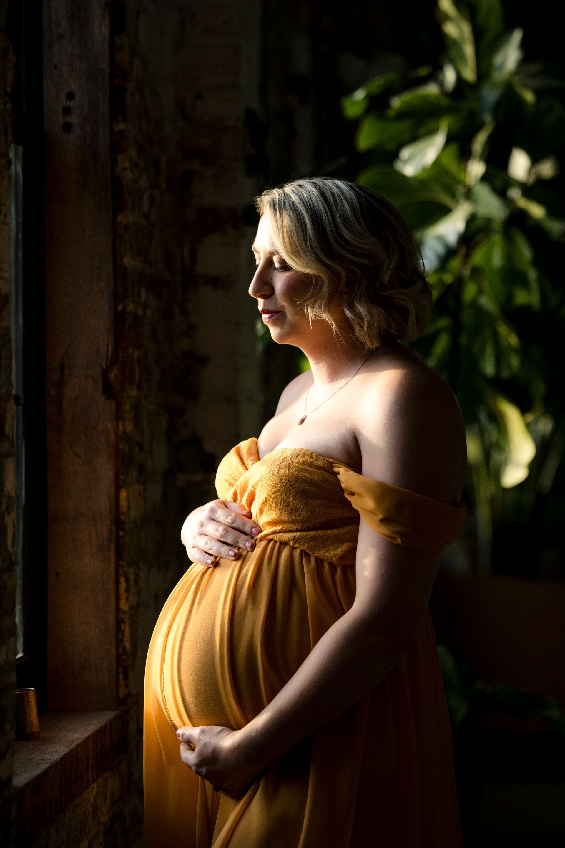 Pregnant person in a mustard dress stands by a window, softly lit in profile, holding their belly