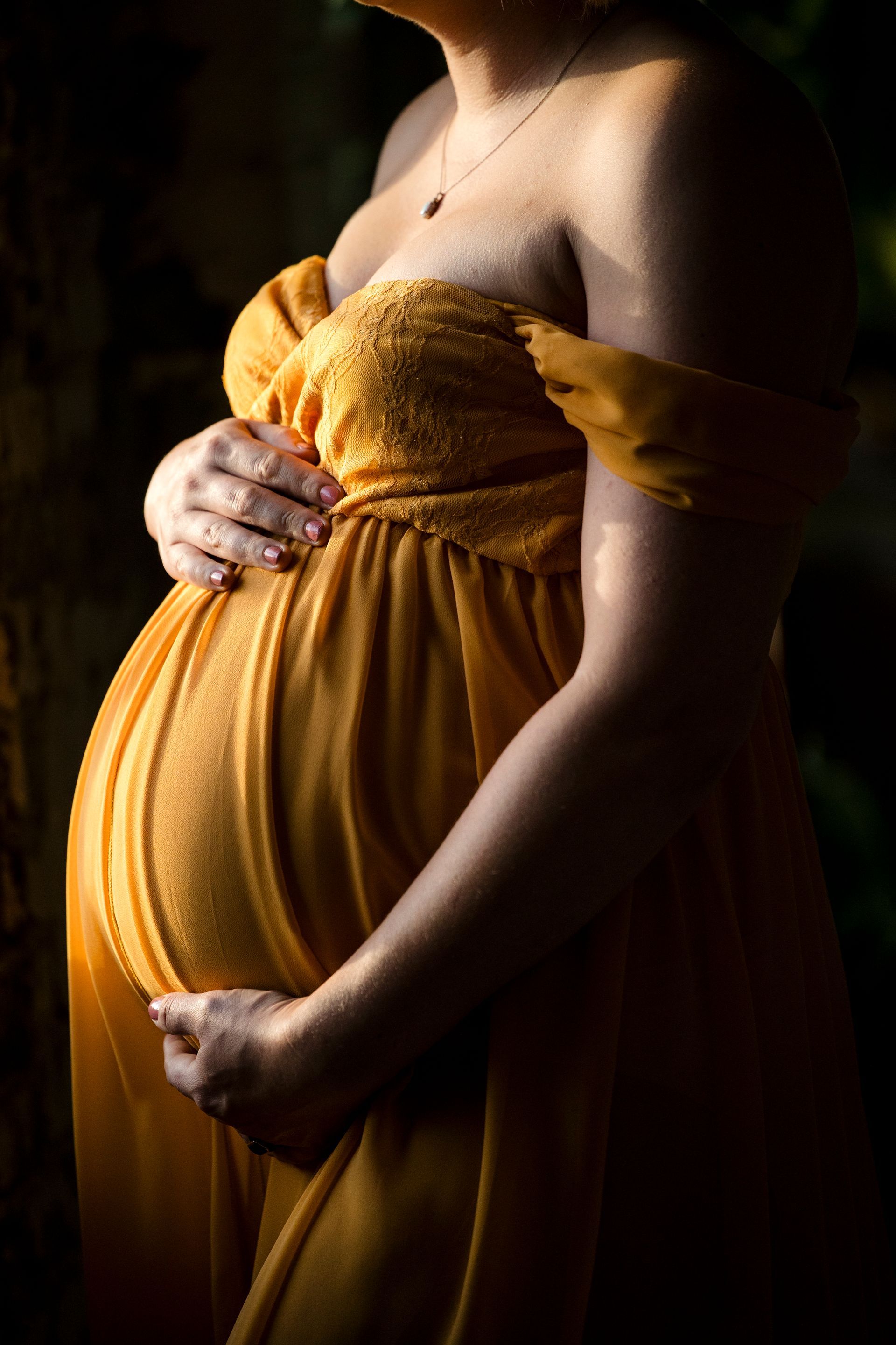 Pregnant person in a mustard-yellow dress holding their belly in soft side light