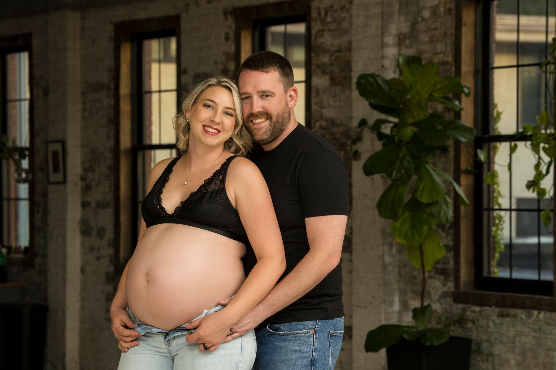 Smiling couple posing together outdoors, with the woman pregnant and the man standing behind her.