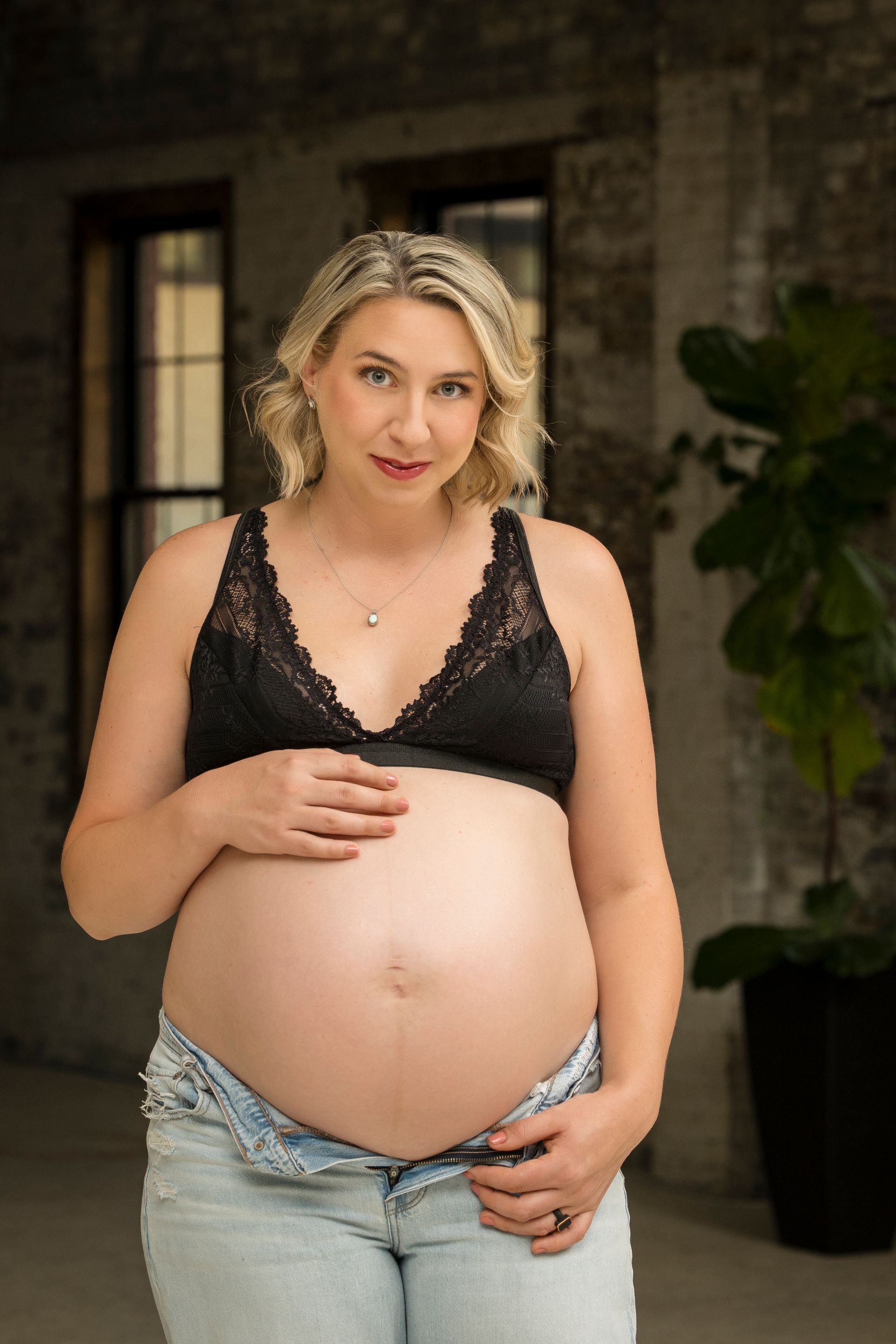 Pregnant person in black top and jeans standing outdoors by a brick wall, hands on belly.
