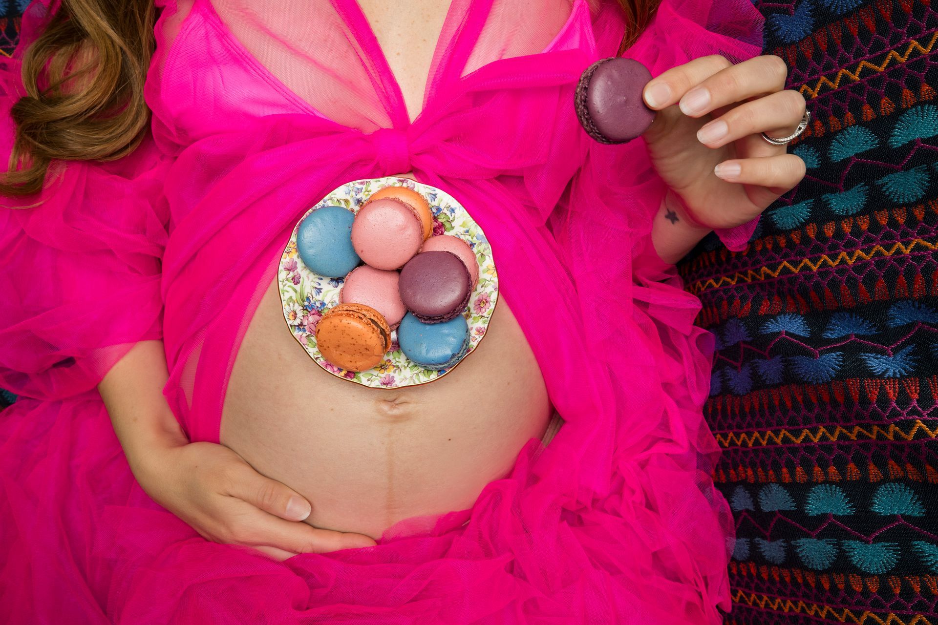 Pregnant belly with colorful macarons arranged like a cupcake top, held by hands, bright pink fabric around it