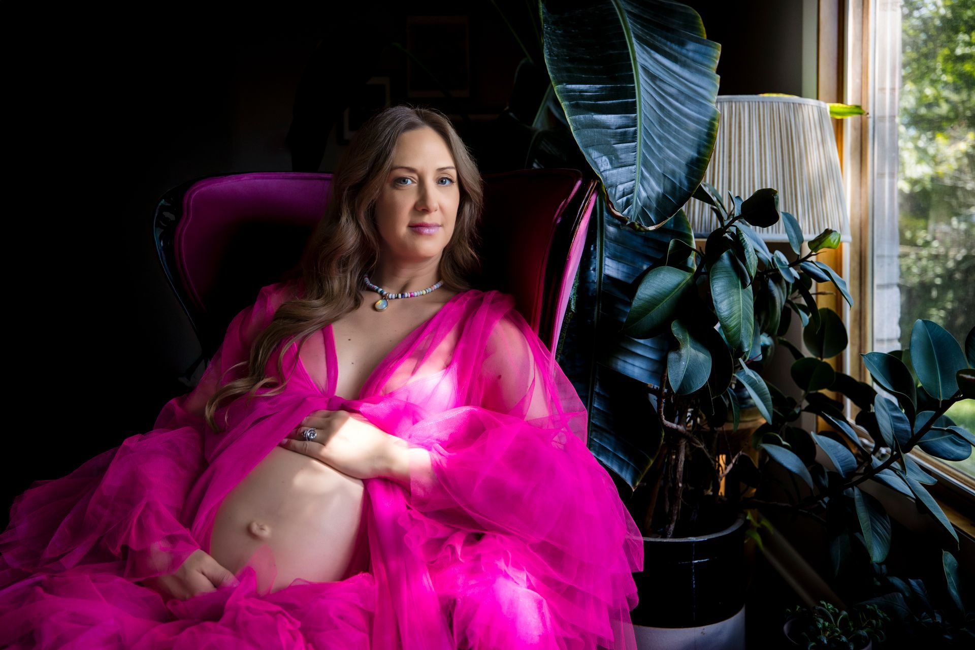 Pregnant person in a bright pink dress reclining by a window with houseplants