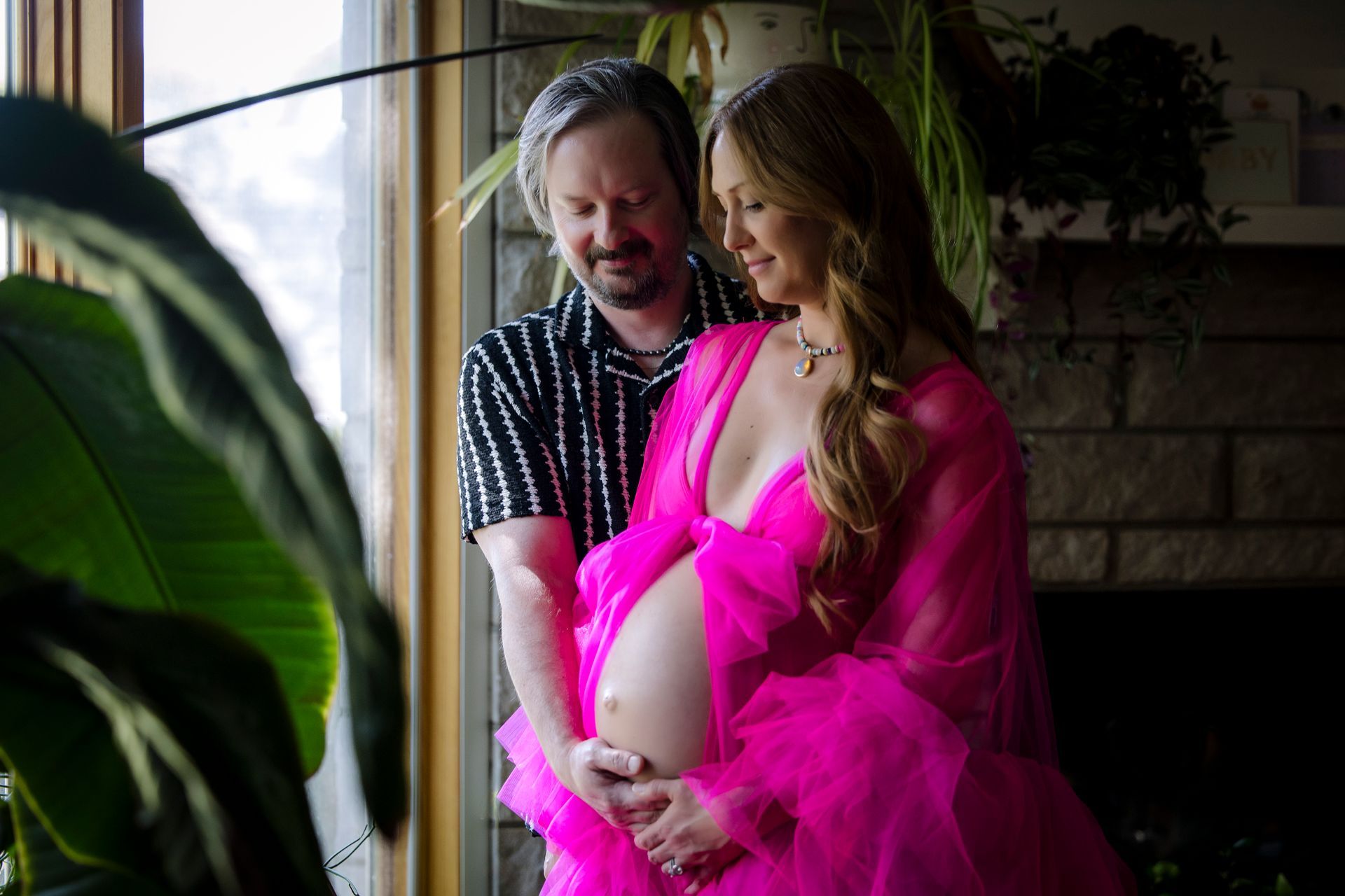 Pregnant woman in bright pink dress with partner by a sunlit window, gently holding her belly