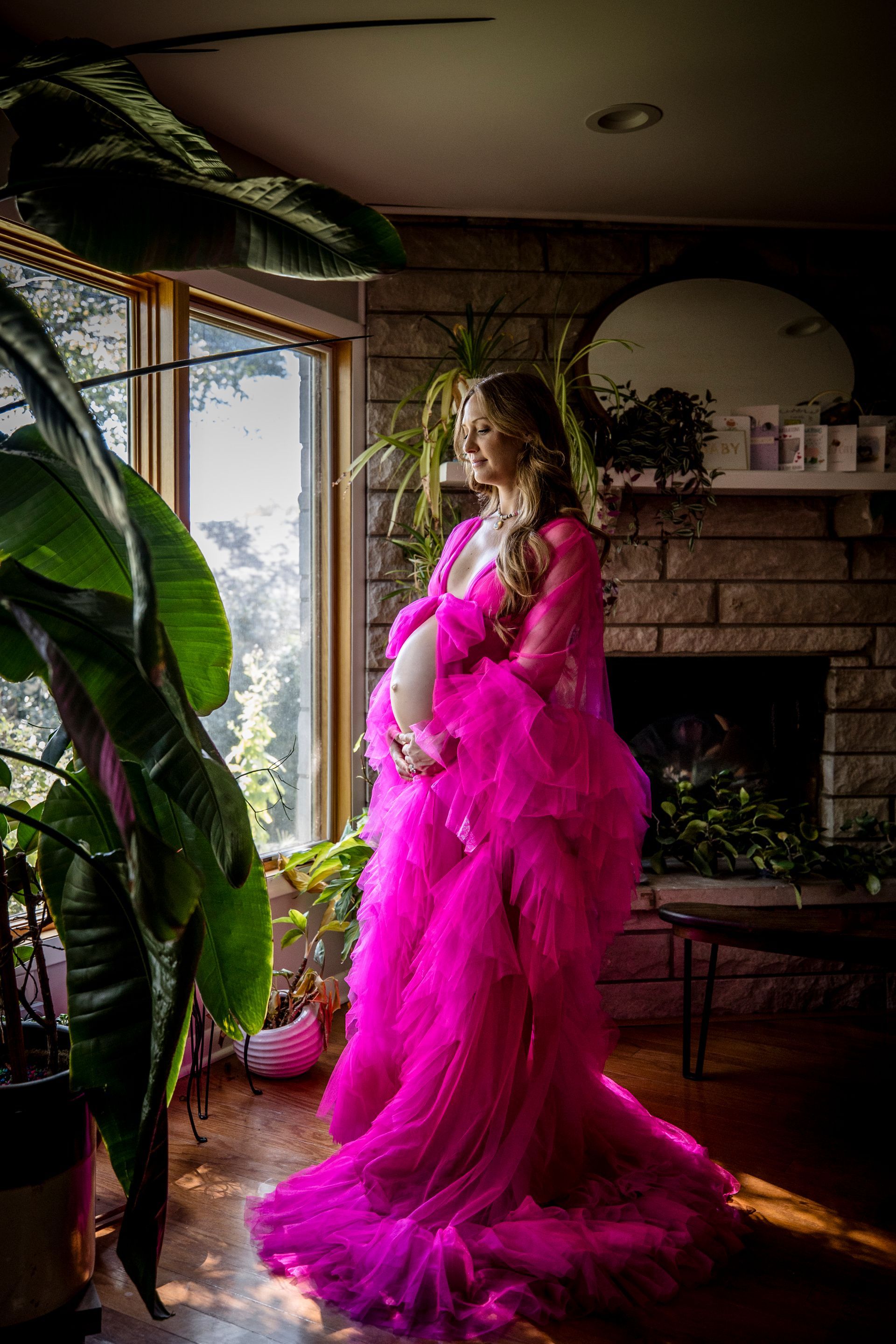 Person in a bright pink feathered gown standing by a sunlit window in a living room