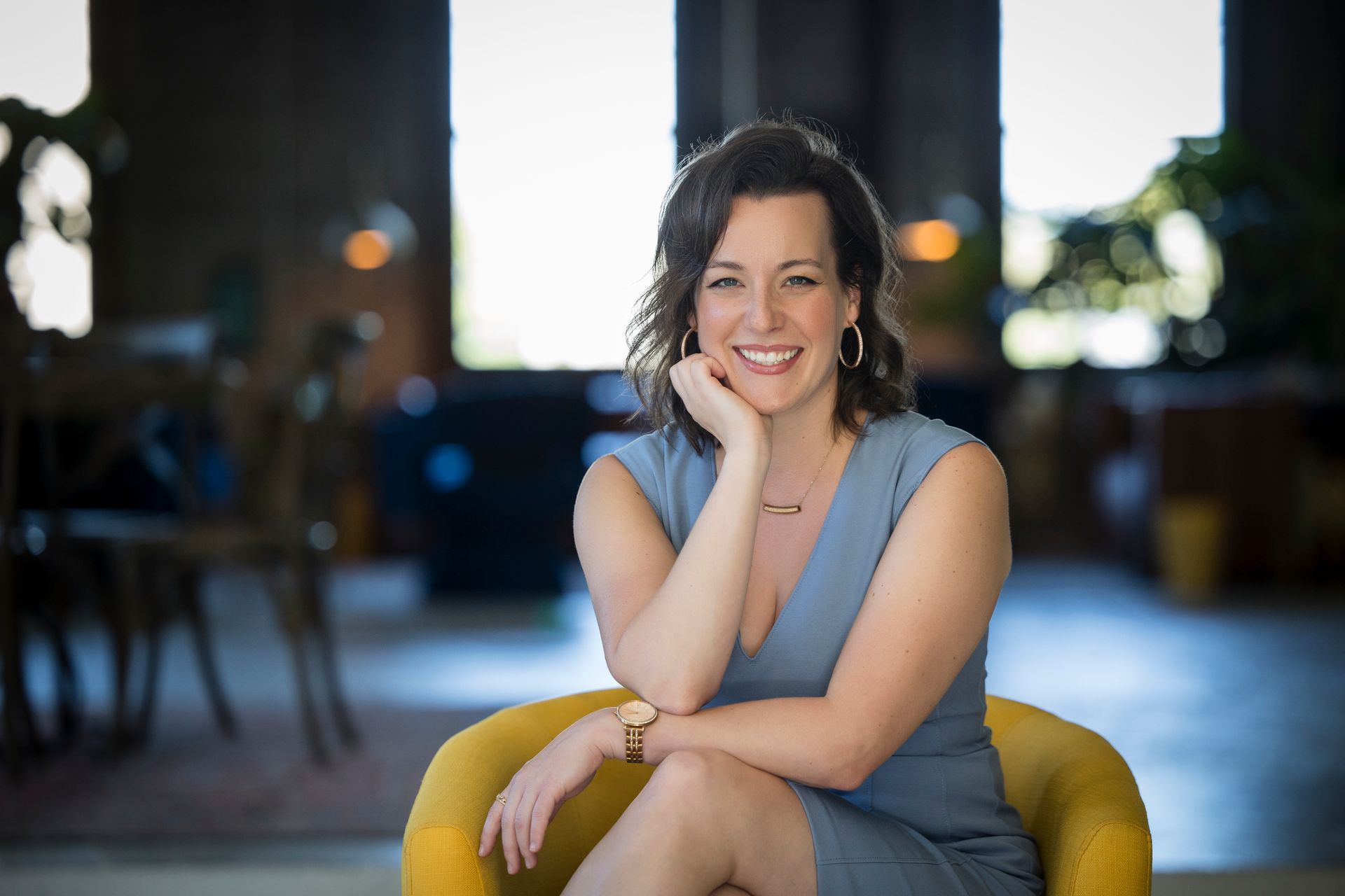 Smiling woman seated on a yellow chair in a softly lit room, resting her chin on her hand.
