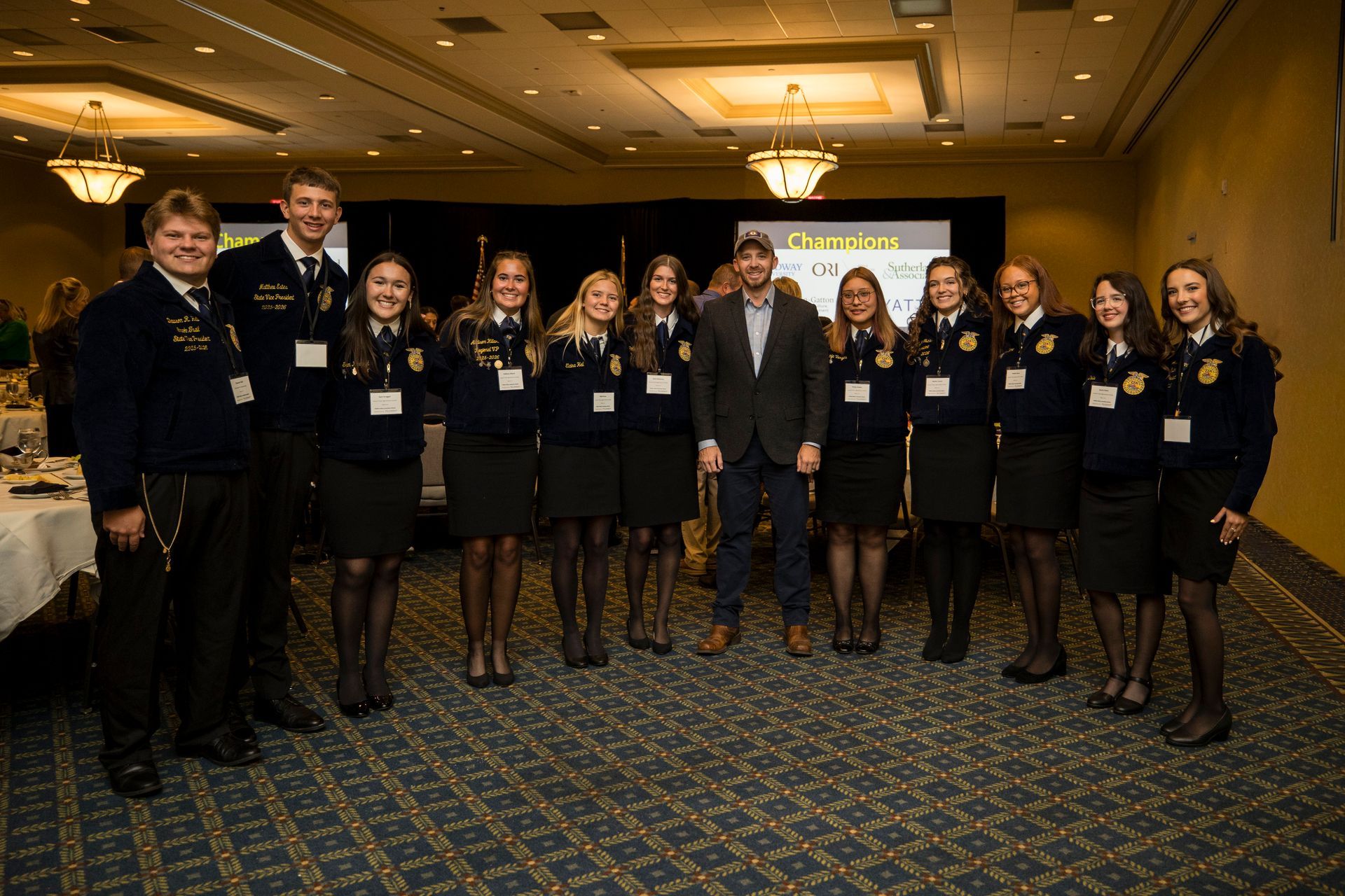Group of people in formal attire posing at an indoor event with a banner in the background.