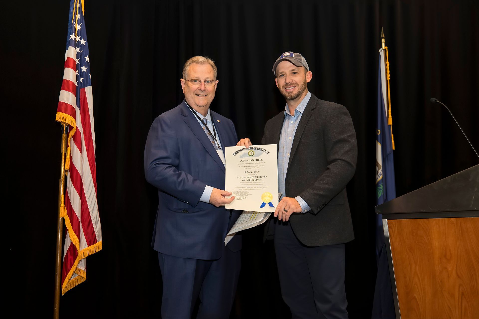 Two men in suits hold a certificate on a stage beside an American flag.