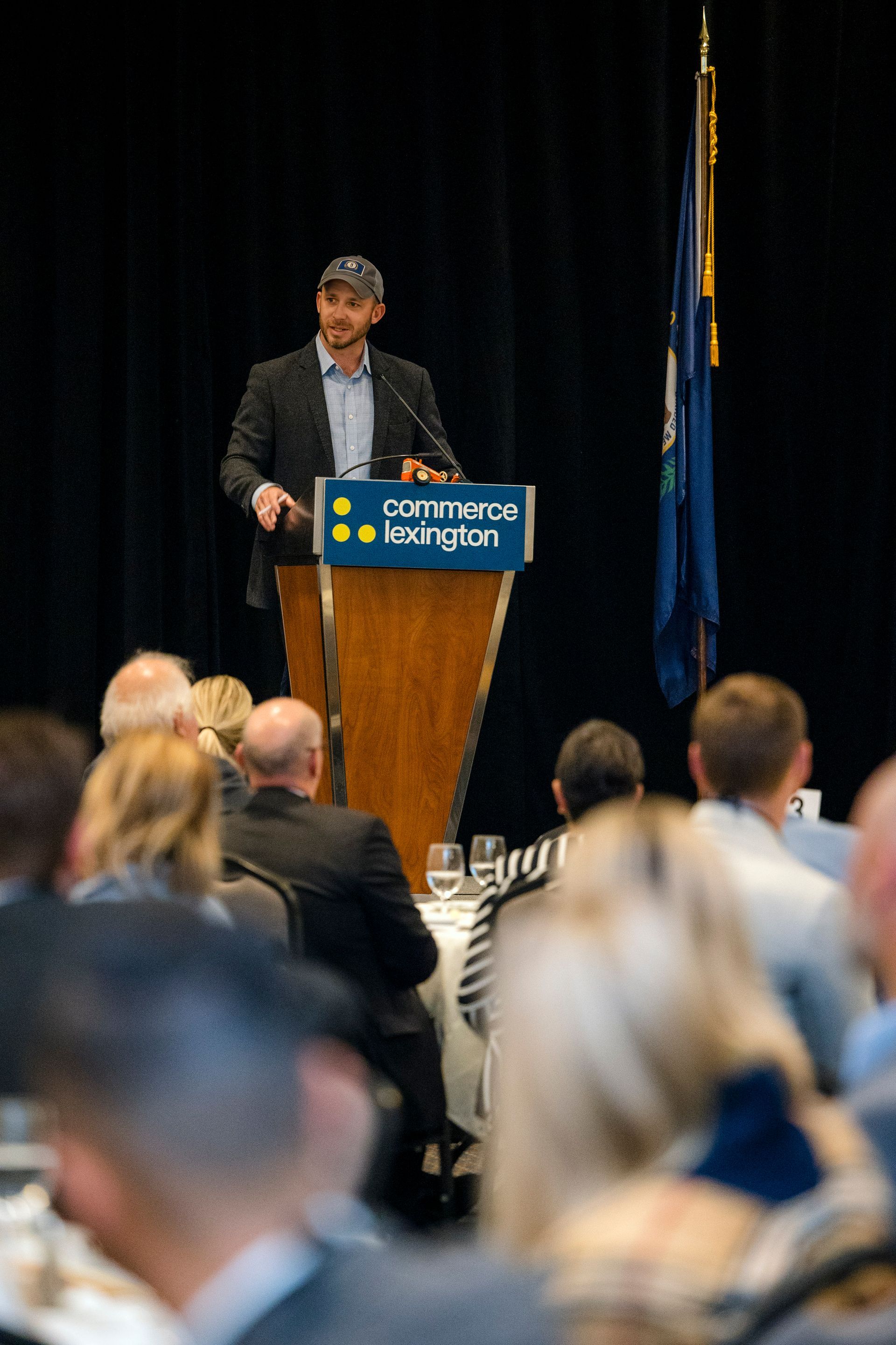Speaker at podium addressing audience in a conference hall, with blurred attendees in foreground.