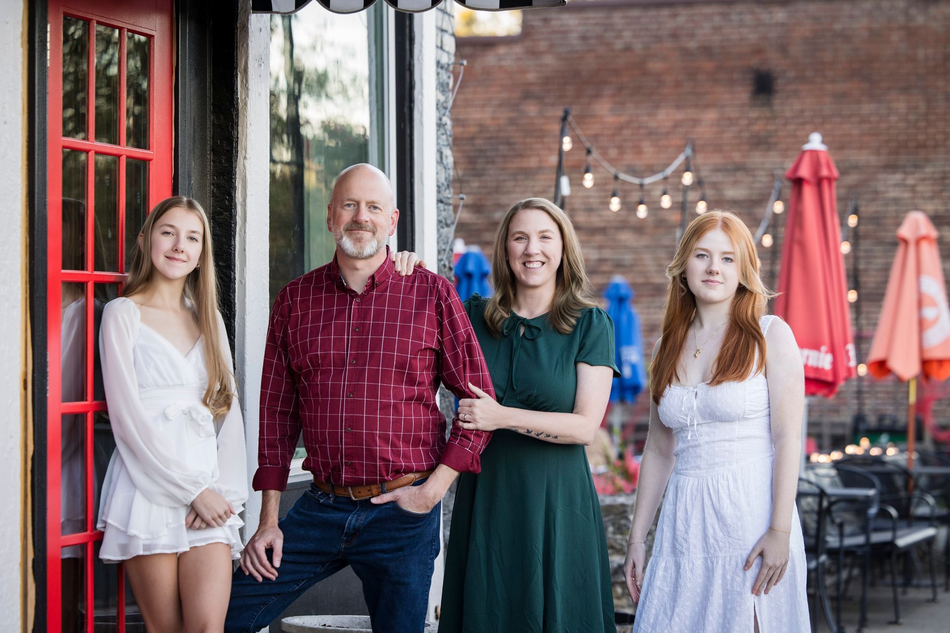 Four people standing outside a restaurant, smiling near red umbrellas and brick walls