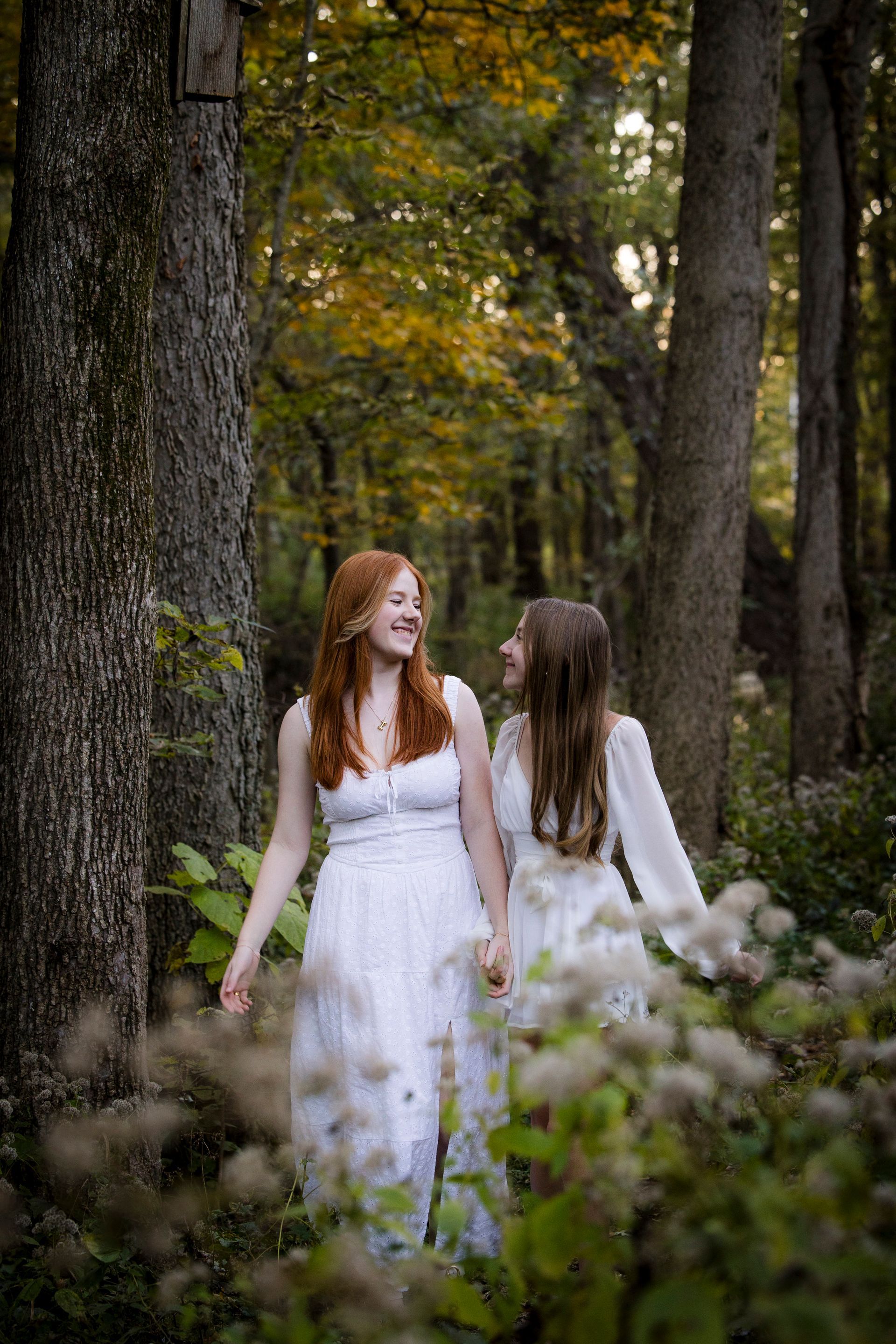 Two women in white dresses walking through a wooded park, holding hands and smiling