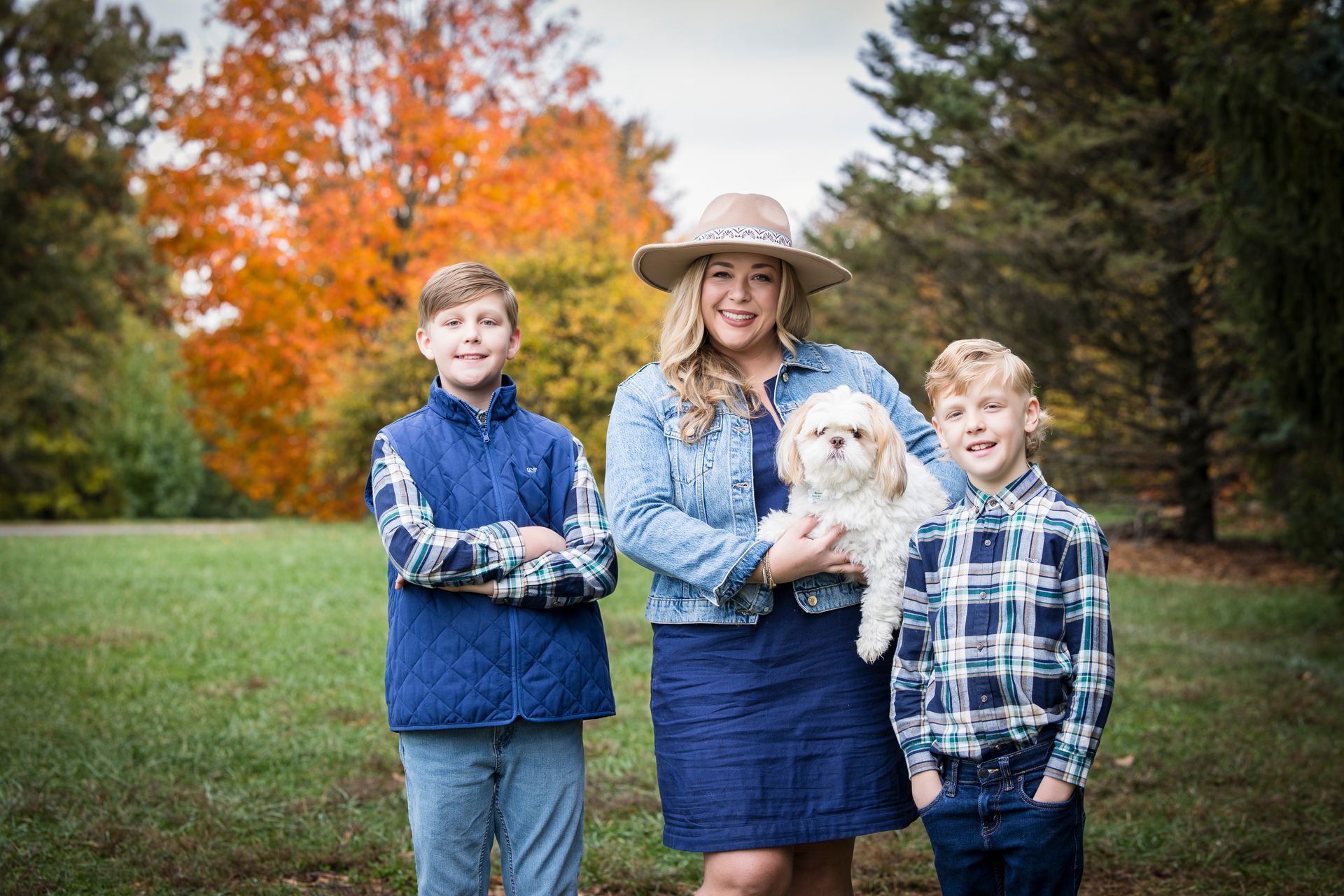 Three people standing outdoors in autumn, holding a small white dog, with colorful trees in the background