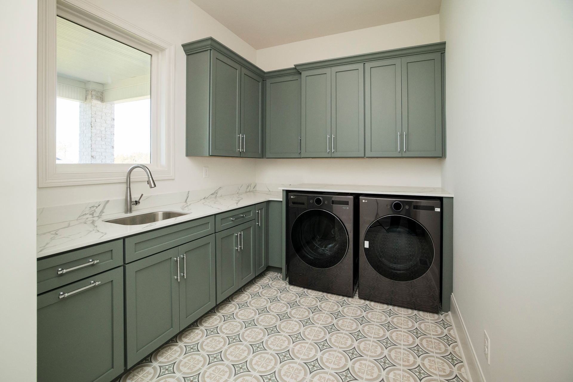 Laundry room with sage green cabinets, white countertops, and front-loading washer and dryer on patterned tile floor