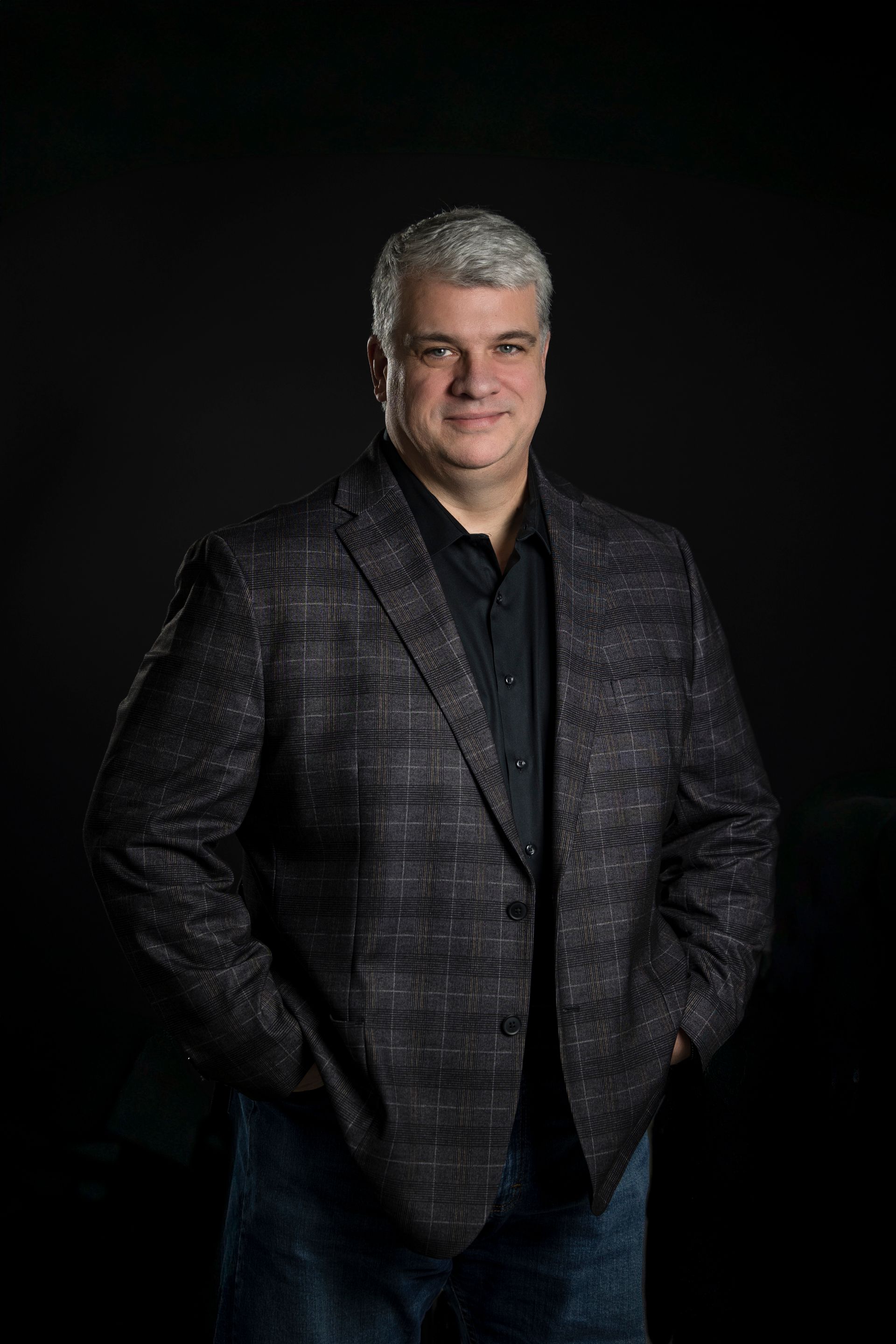 Studio portrait of a smiling man in a dark plaid blazer against a black background