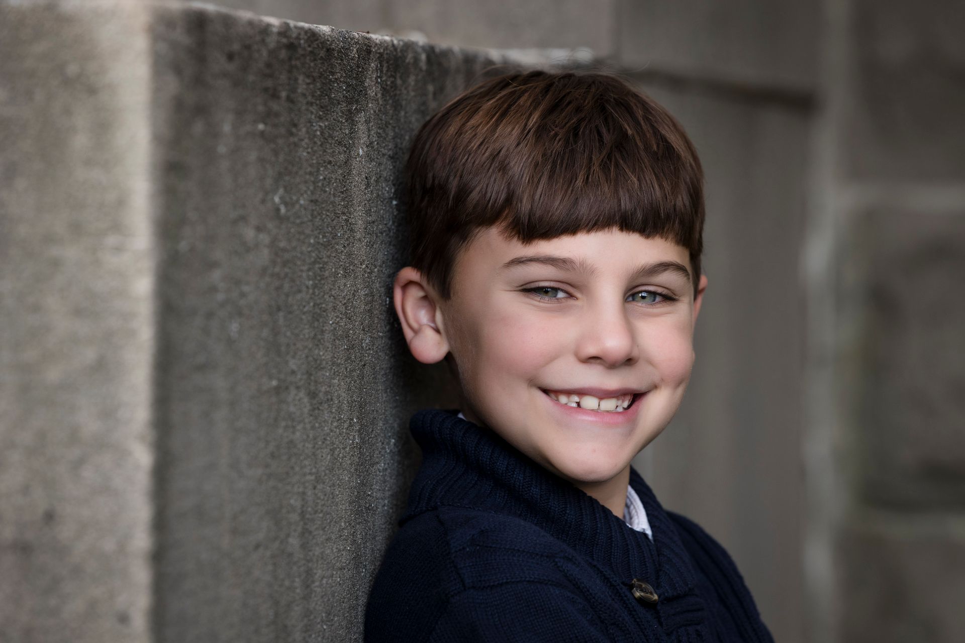 Smiling child in a navy sweater leaning against a concrete wall