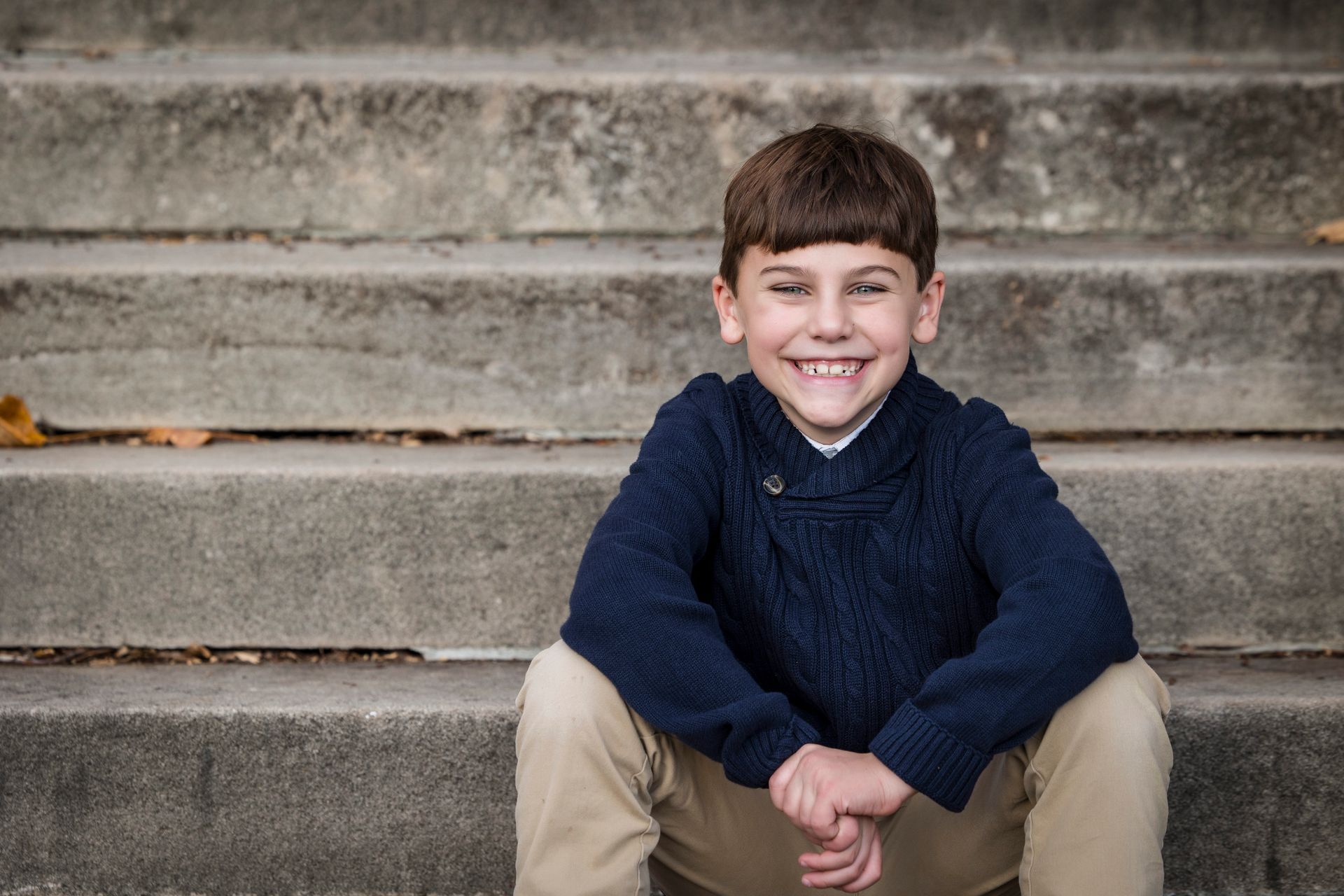 Smiling child sitting on stone steps, wearing a navy sweater and beige pants.