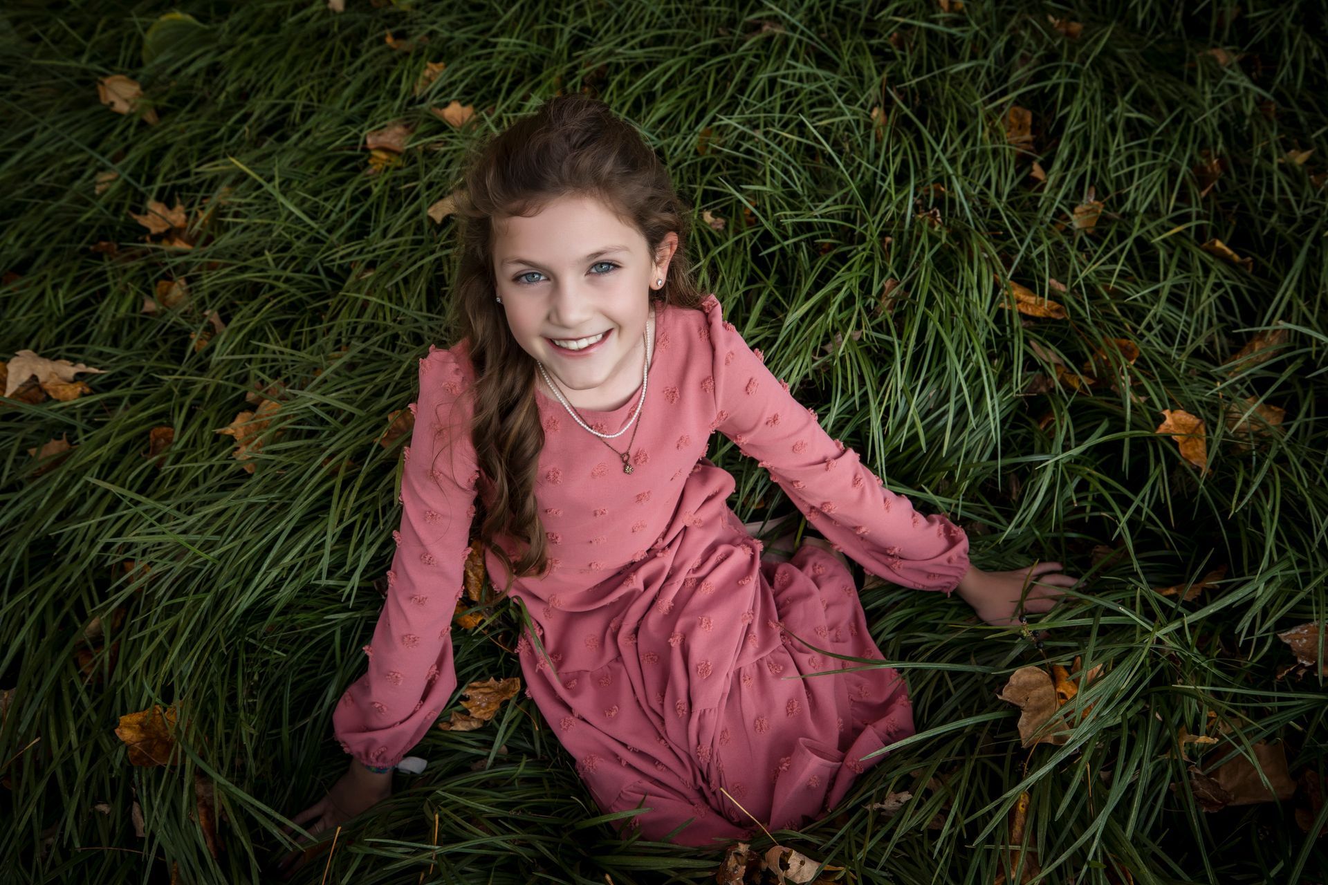 Smiling child in a pink dress lying in tall grass with autumn leaves