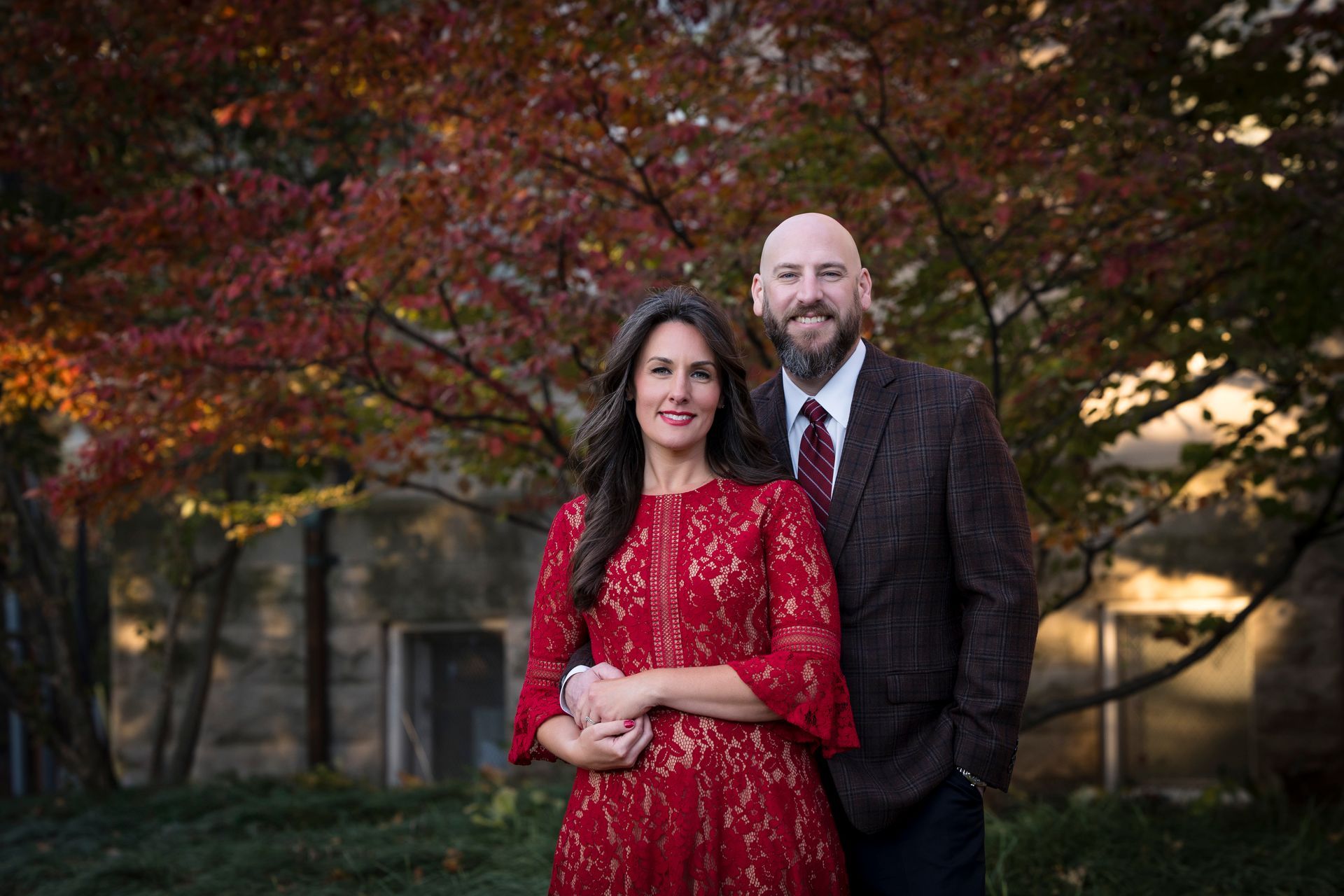 Couple standing outdoors in formal attire, with autumn trees in the background.