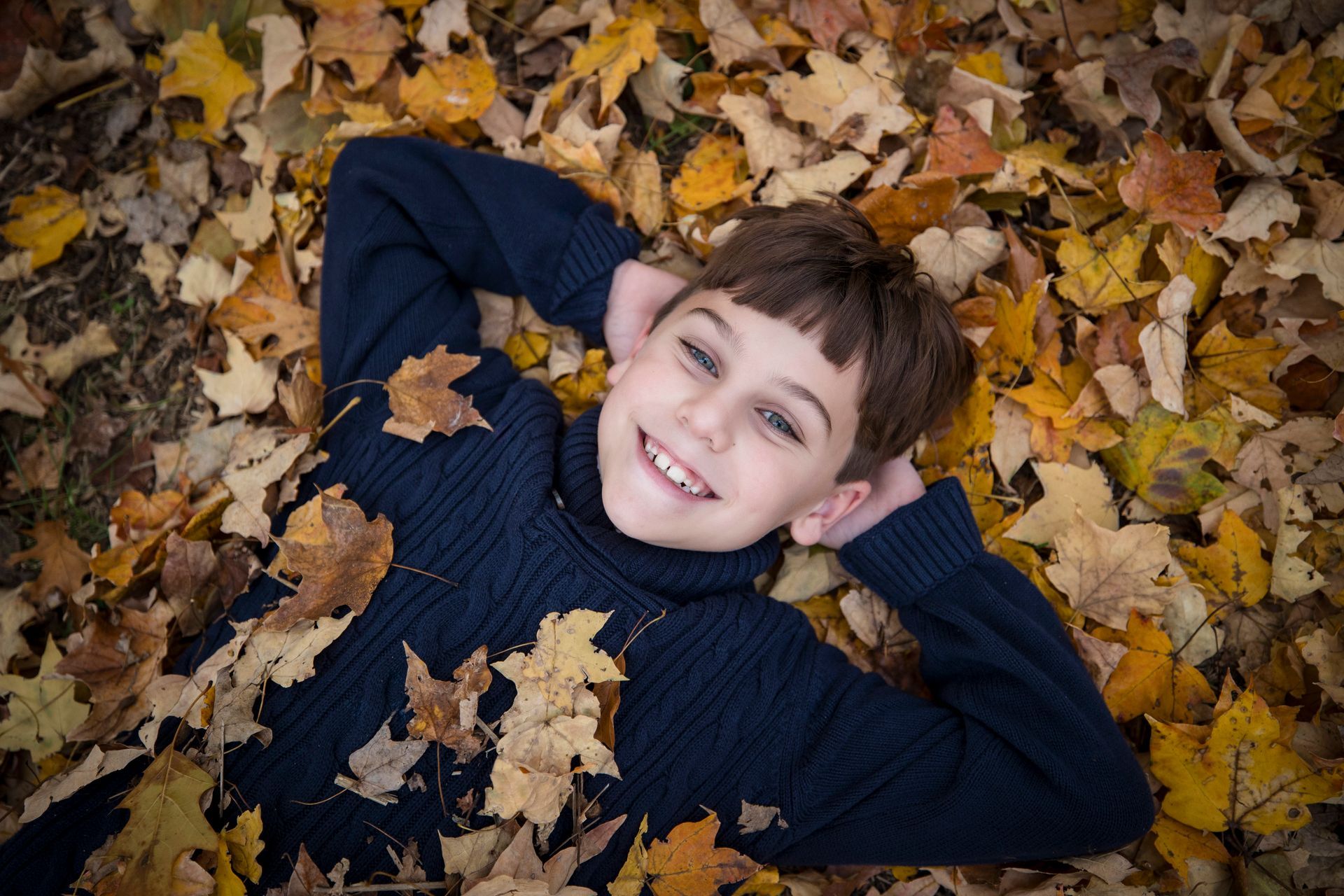 Smiling child lying in autumn leaves, wearing a dark sweater.