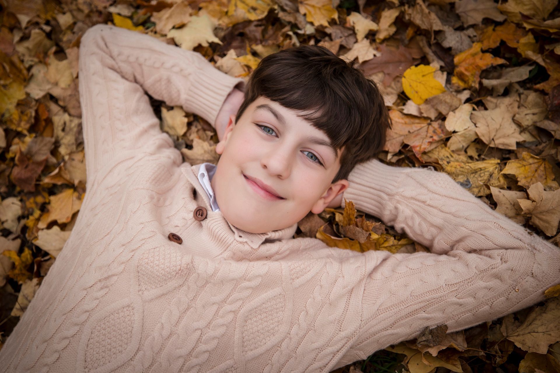 Smiling person lying on autumn leaves with hands behind head, wearing a beige sweater
