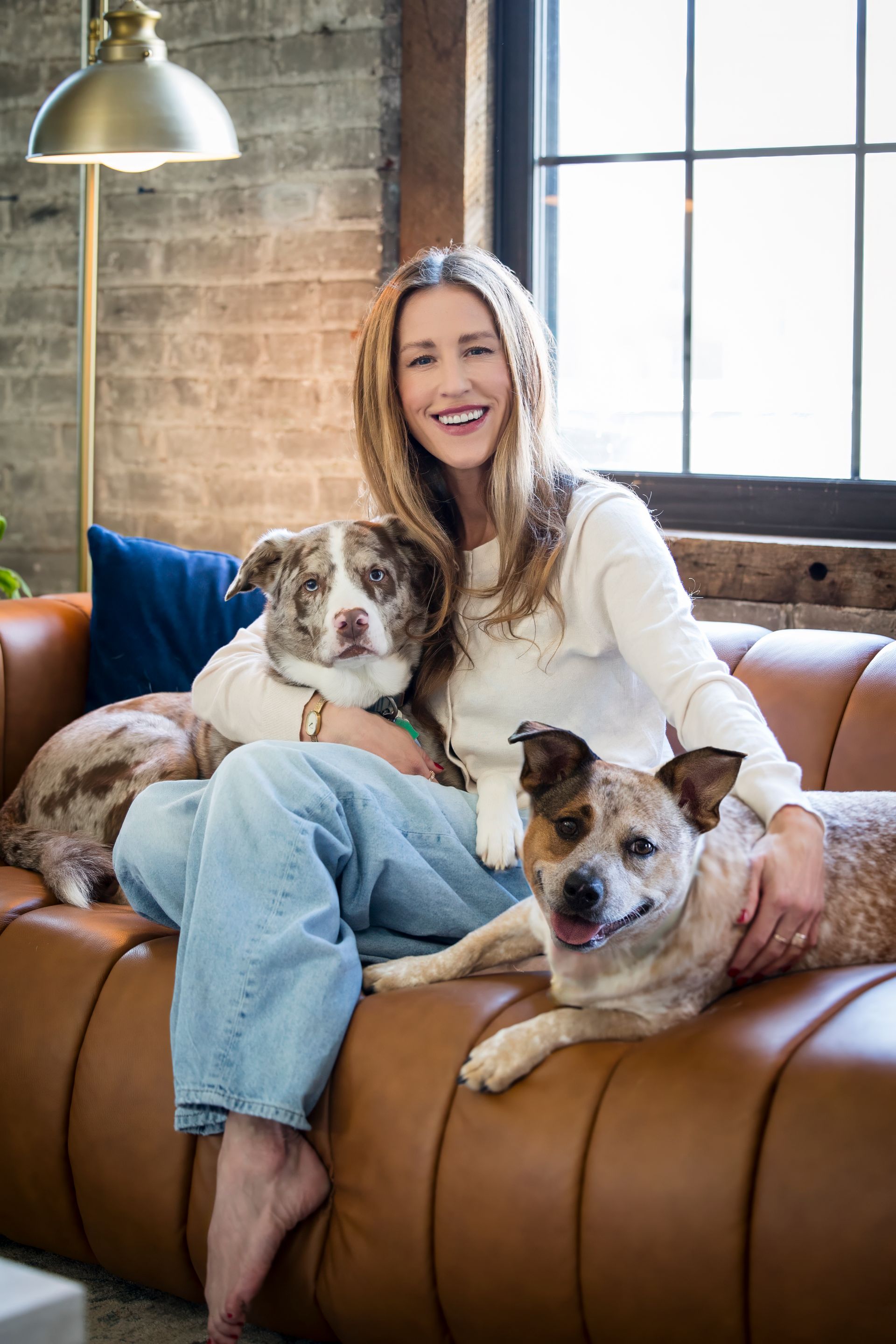 Woman sitting on a tan sofa with two dogs in a sunlit room, smiling at the camera.