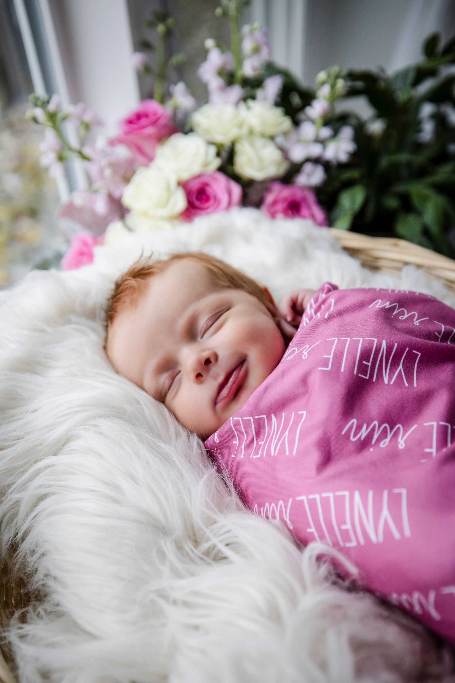Sleeping baby wrapped in pink blanket on a fluffy white bed with flowers in the background