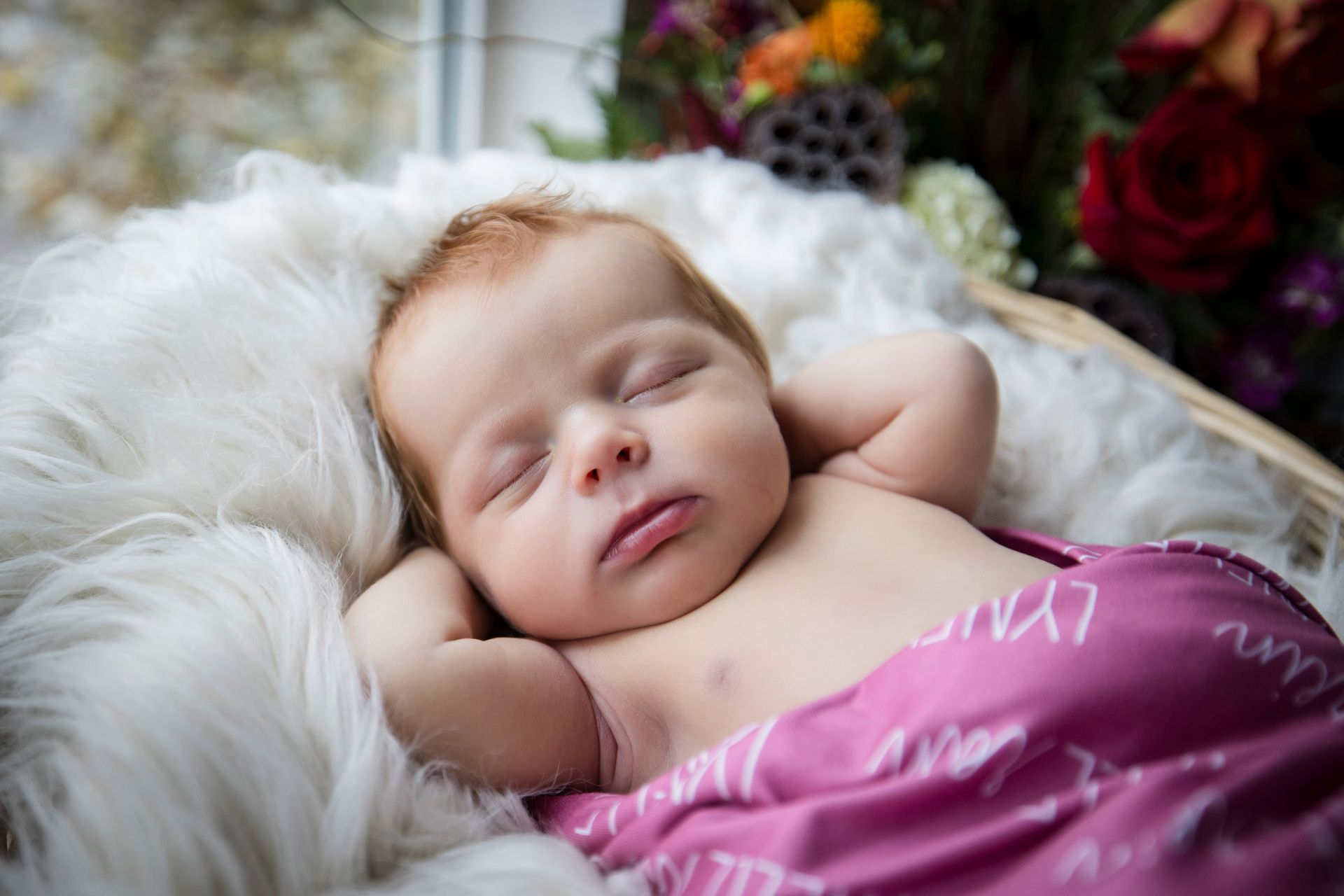 Sleeping newborn wrapped in a pink blanket, resting on a fluffy white fur backdrop with flowers nearby
