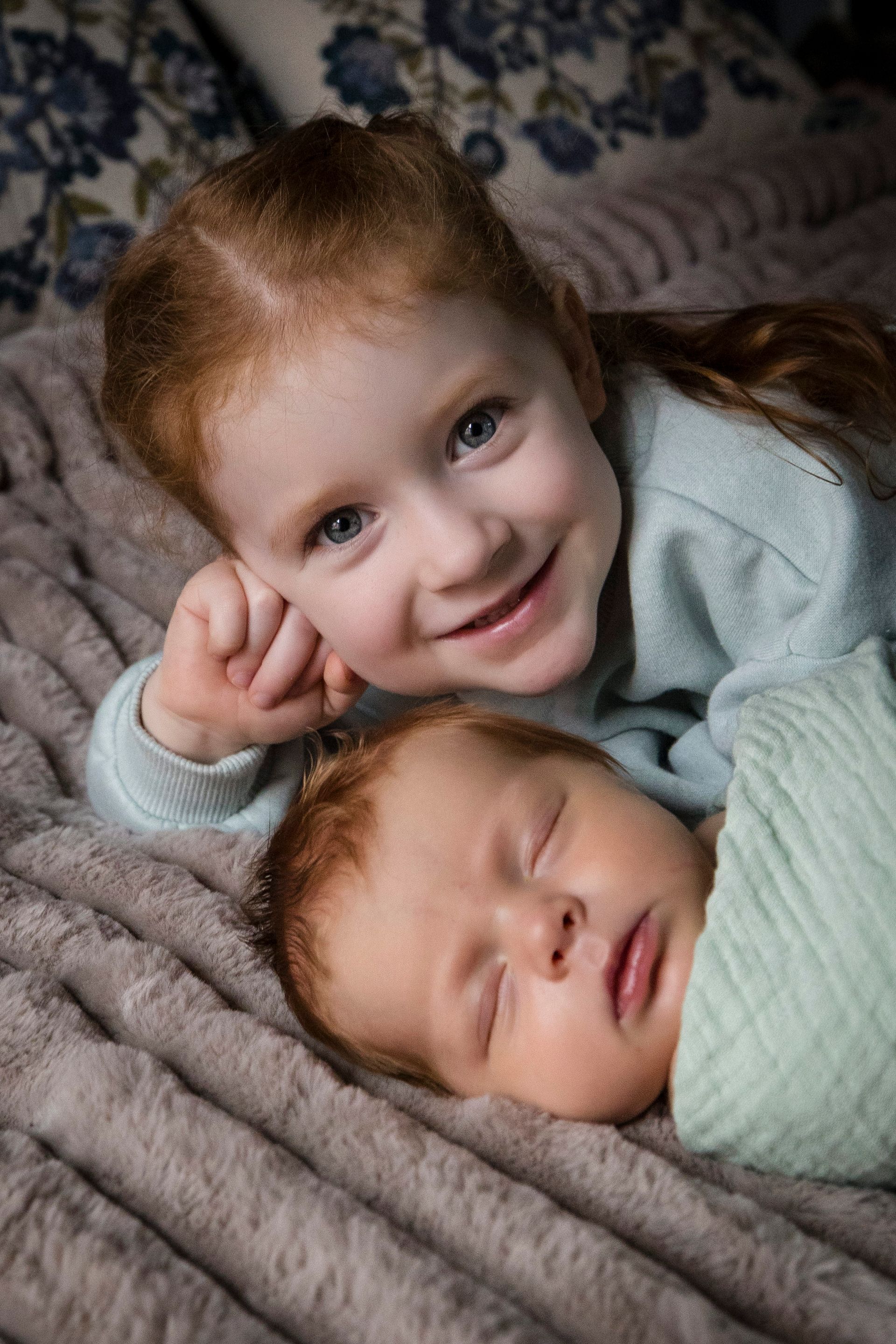 Two children lying together on a soft gray blanket, one smiling at the camera.