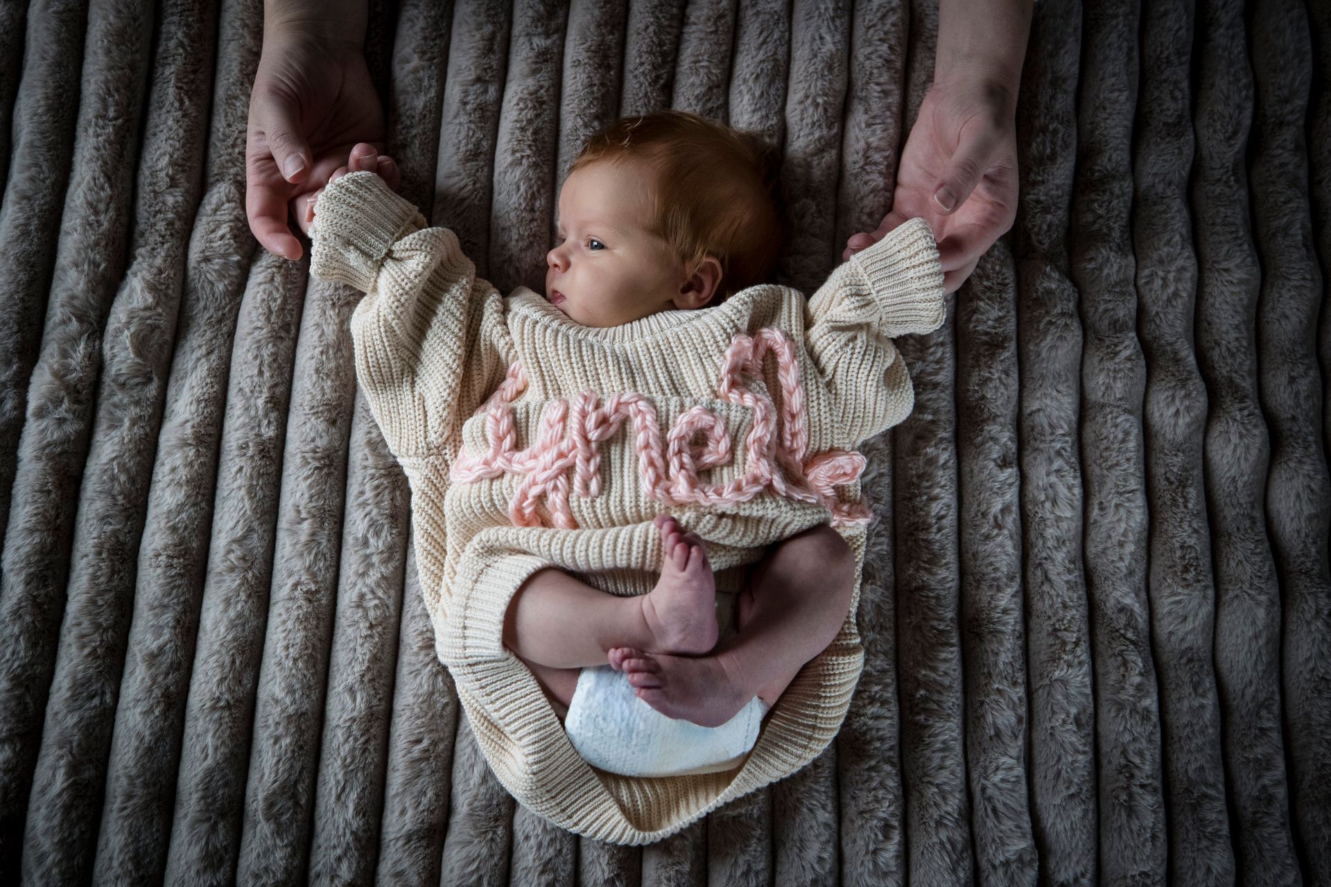 Baby in a cream knit outfit lying on a gray textured blanket, arms held up by adult hands.