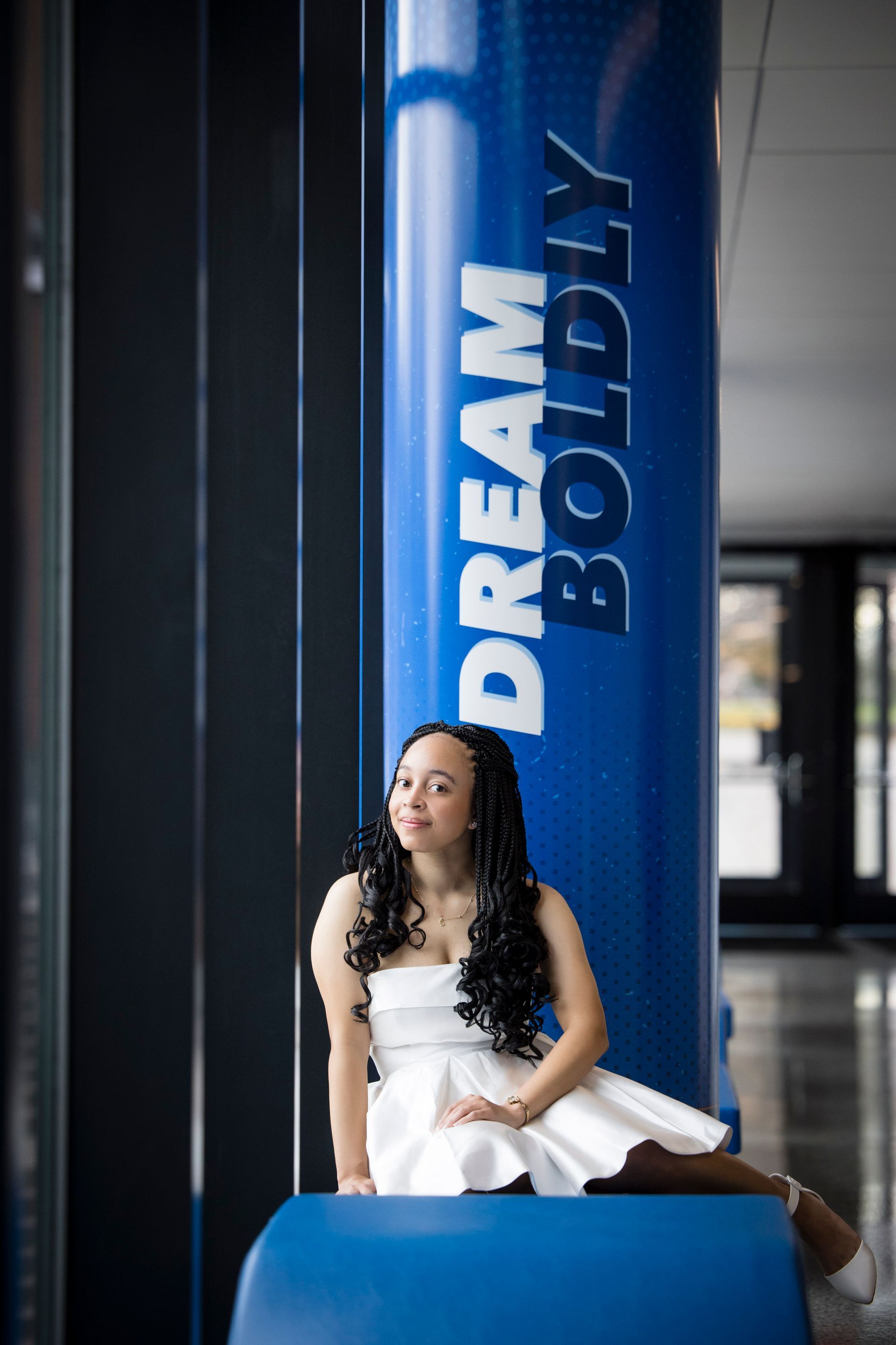 Woman in a white dress sitting beside a blue “DREAM BIG” pillar in a modern interior.