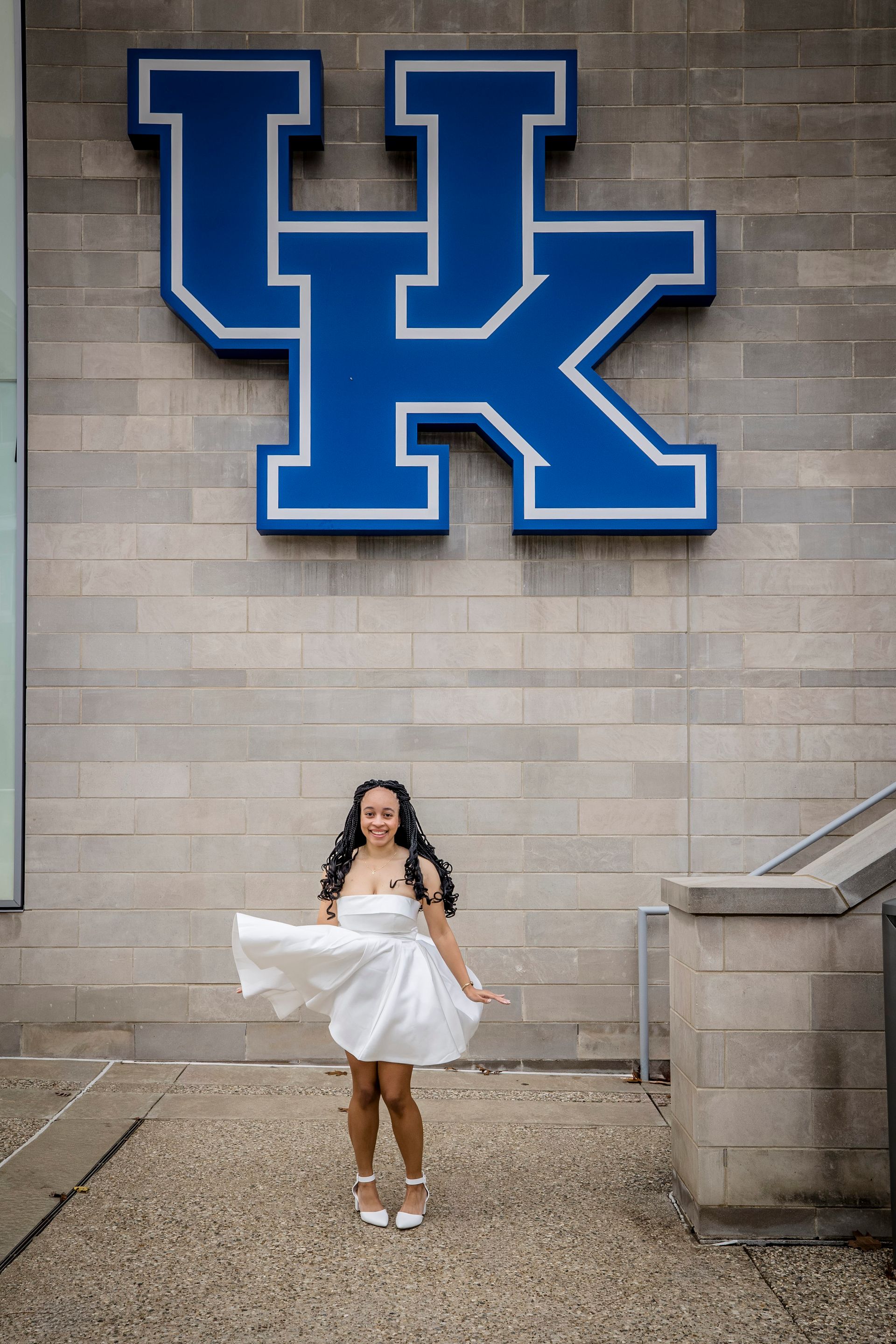 Woman in a white dress posing beneath a large blue UK logo on a brick wall