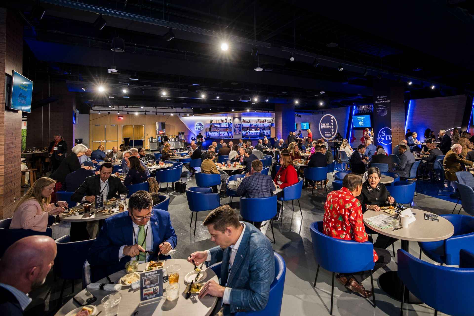 Crowded banquet hall with people seated at round tables under blue lights, watching a stage at the front