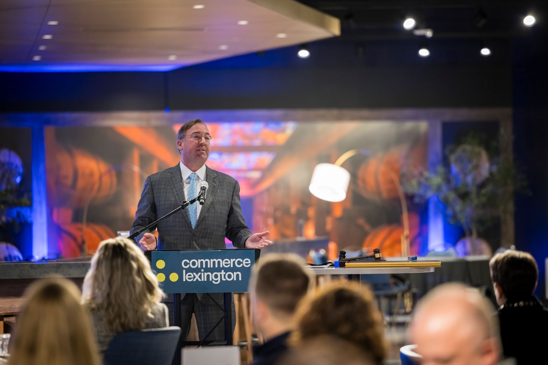 Speaker at podium on conference stage with audience, colorful backdrop, and a lit lamp behind him
