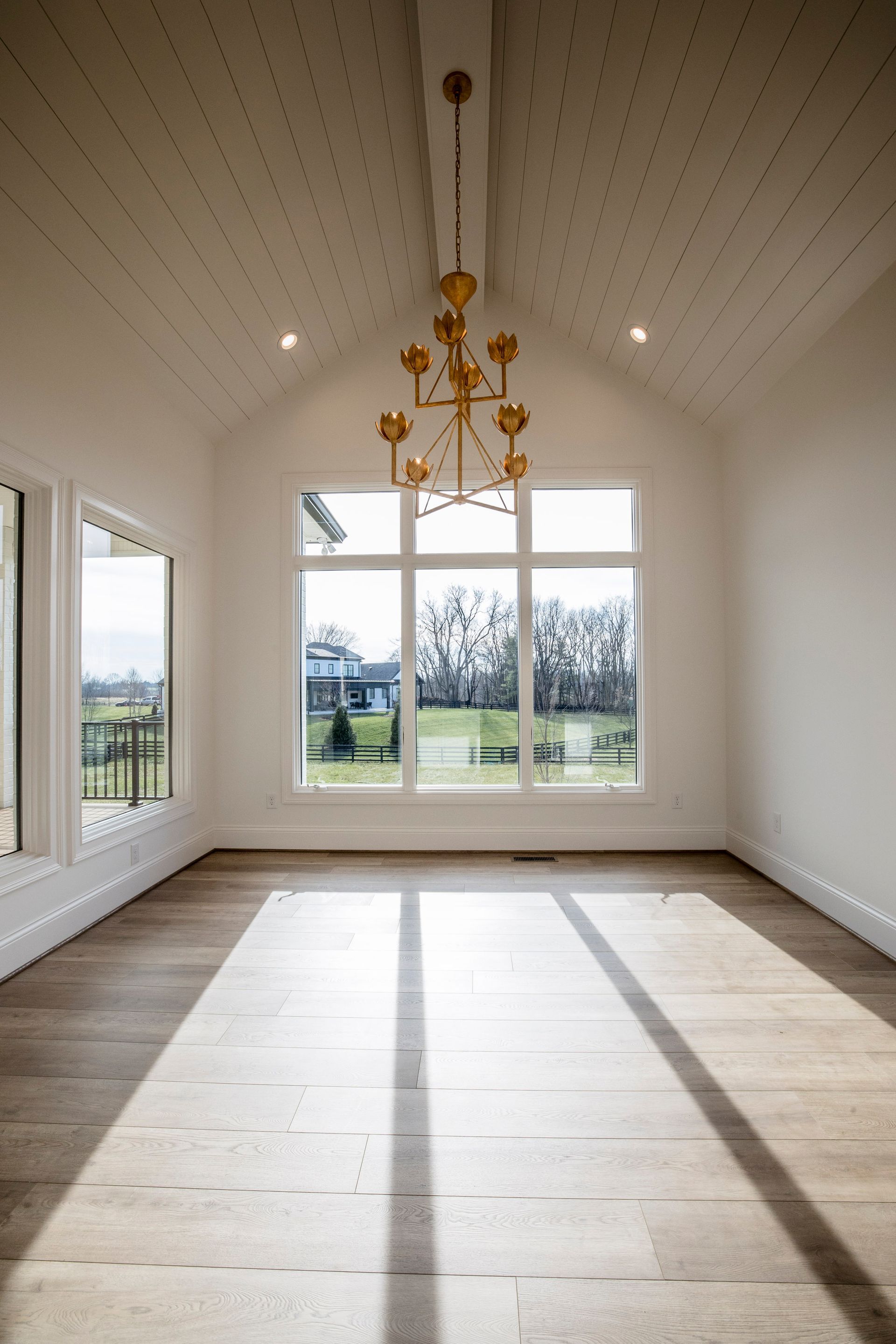 Bright empty room with vaulted ceiling, large windows, and sunlight on the wooden floor.