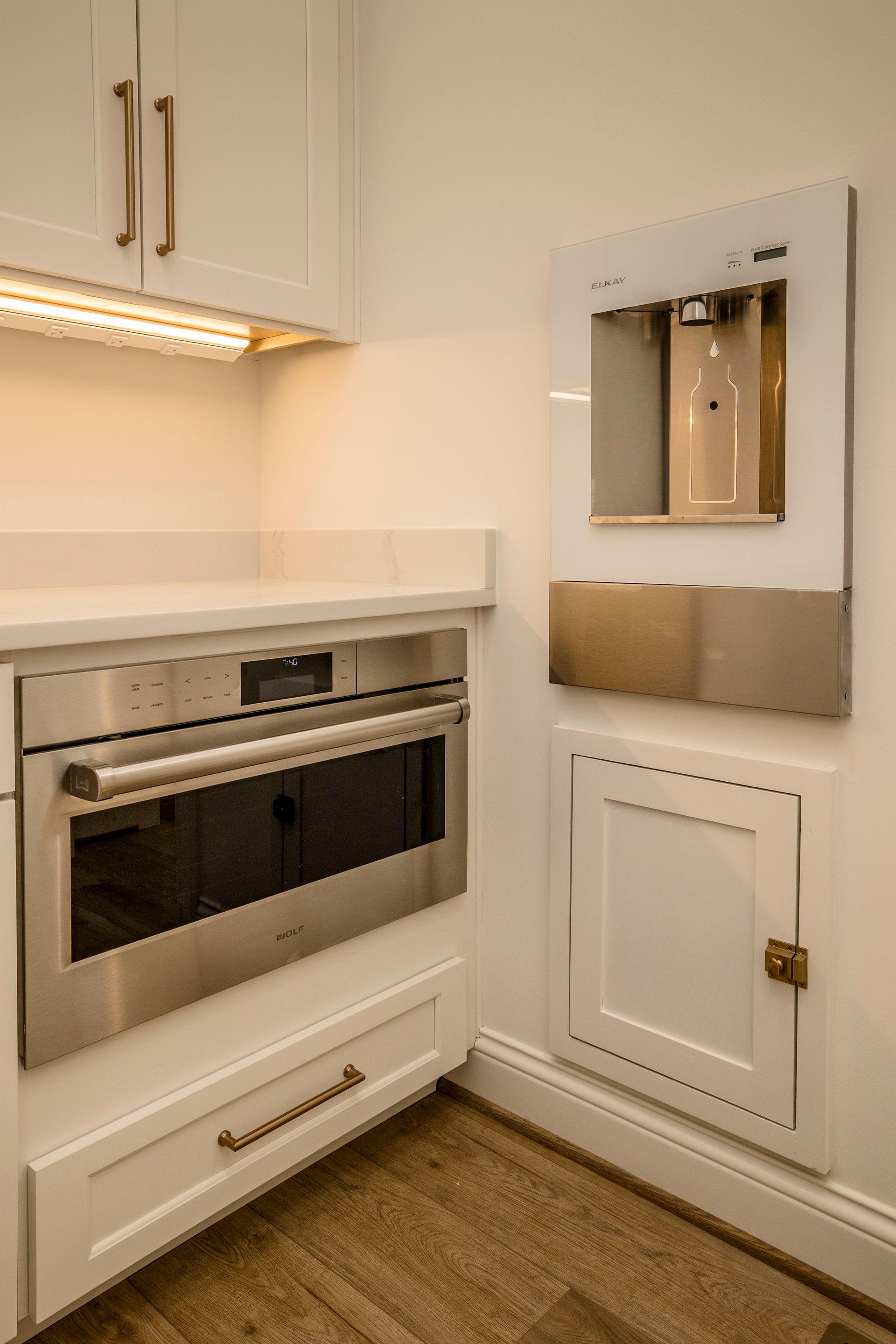 White kitchen corner with built-in oven, cabinets, and a wall mirror above a small door panel.