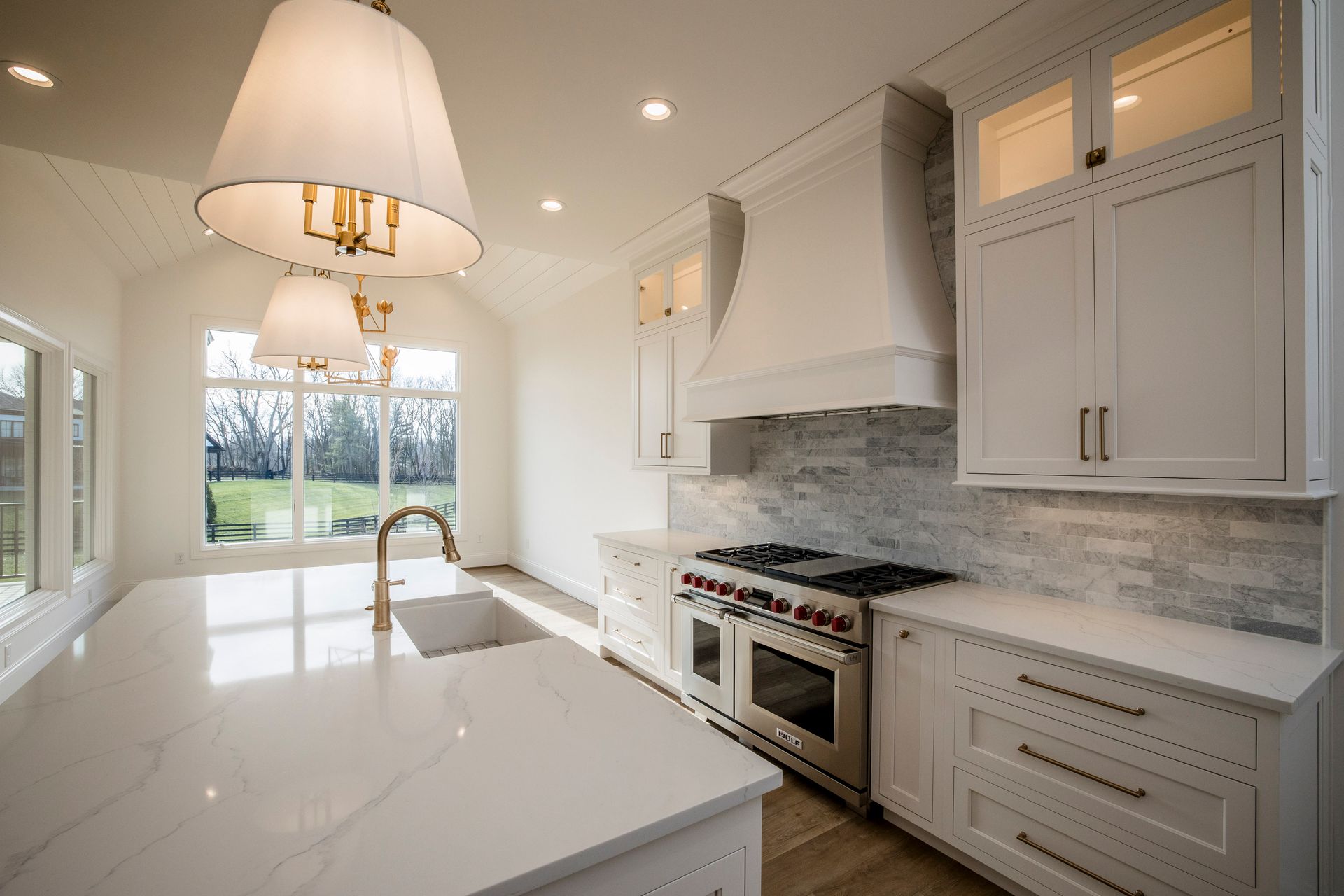 Bright modern white kitchen with island, pendant light, marble counters, and large window to backyard.