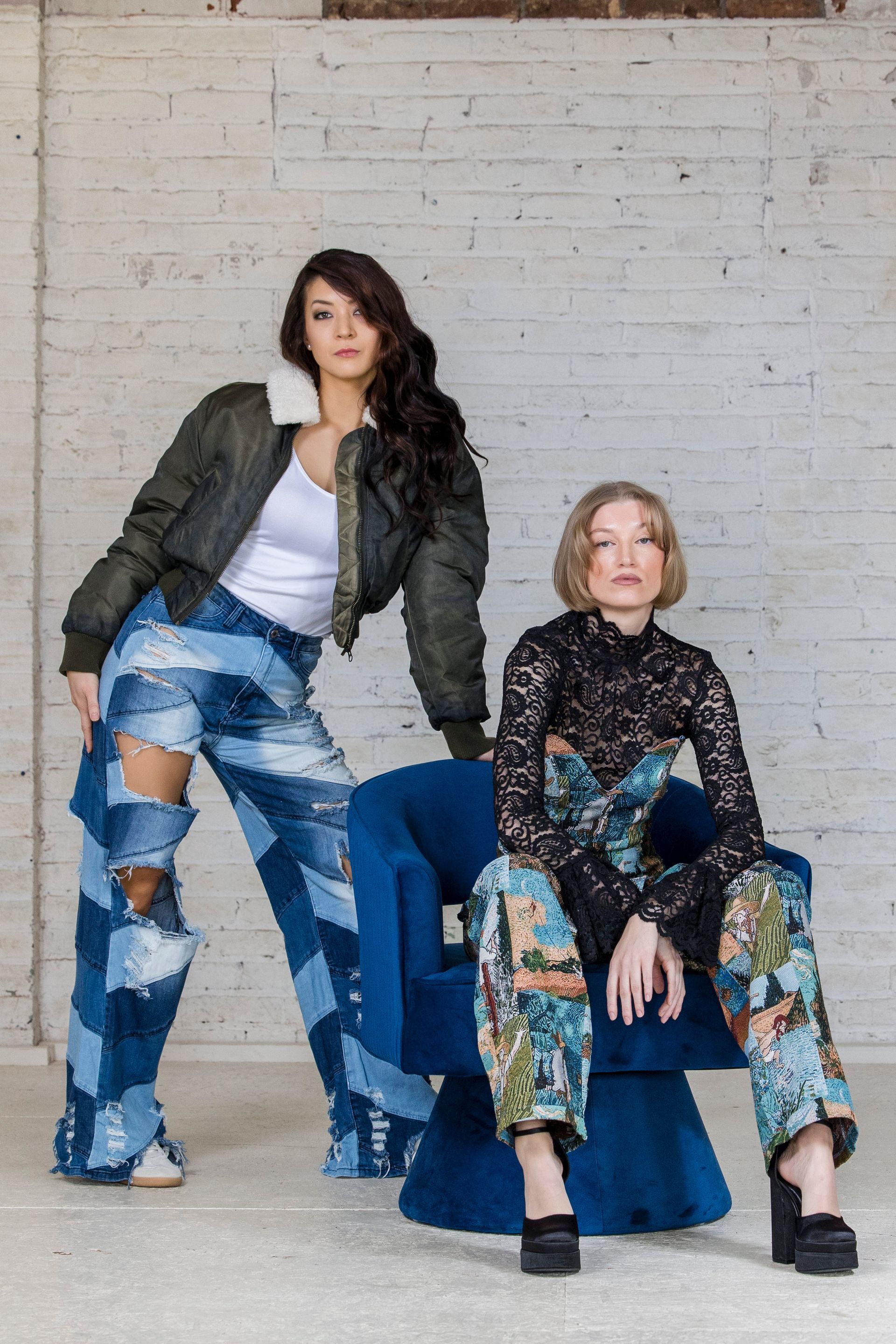 Two women pose in fashion-forward outfits against a white brick wall, one seated on a blue cube.