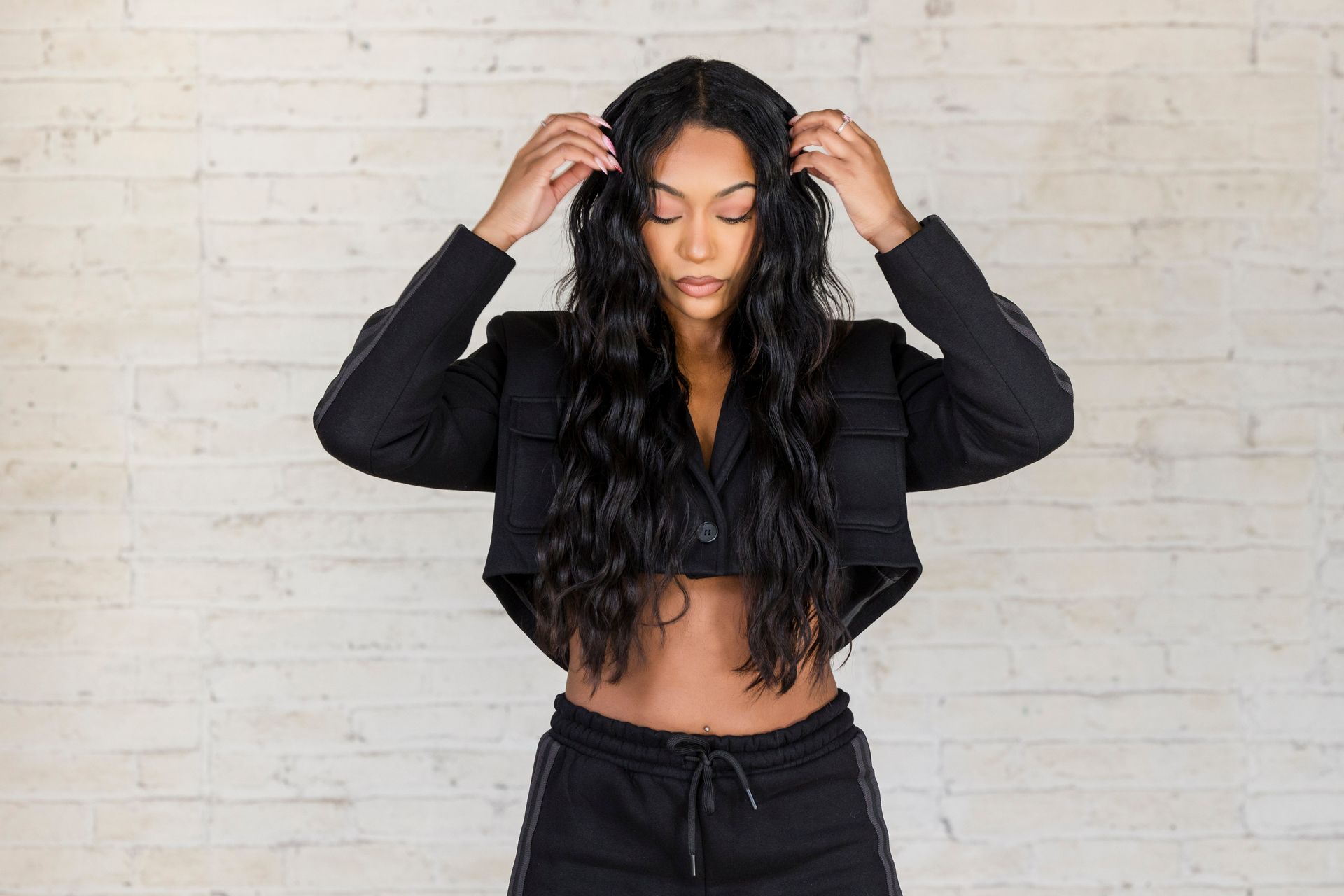 Person with long wavy black hair standing against a white brick wall, adjusting their hair.